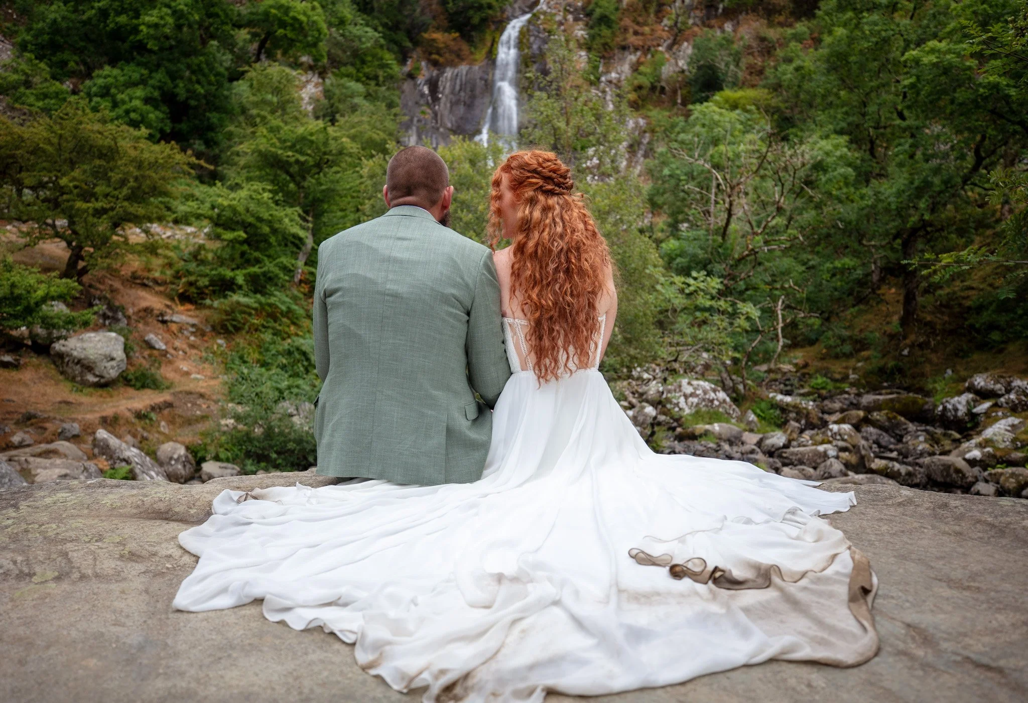 A couple sitting on rocks facing a waterfall, the bride's long dress spread out