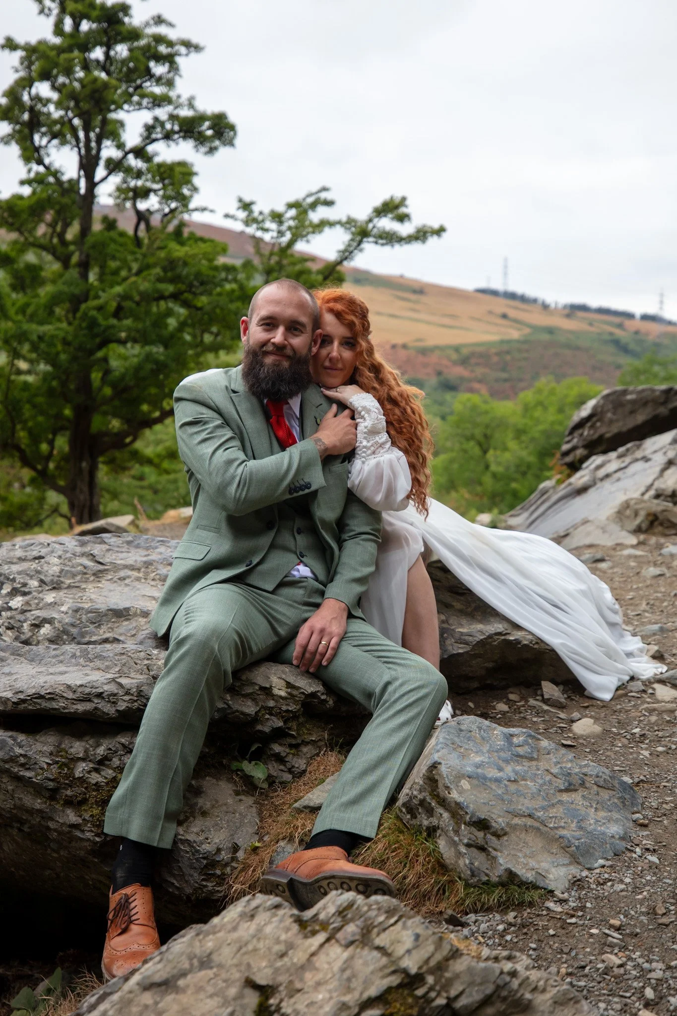Couple sitting on a rock, embracing, with trees and hills behind