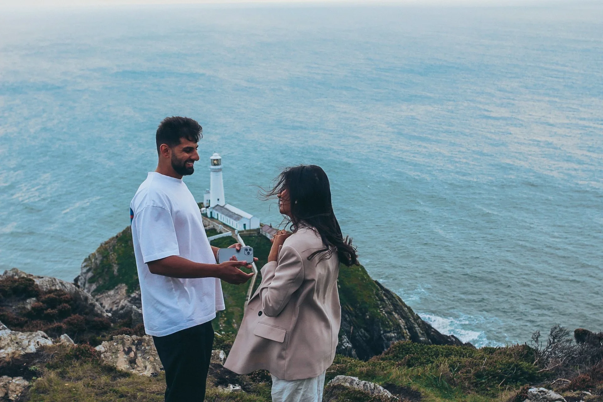 A man and woman sharing a moment on a clifftop above the sea, with a white lighthouse on the rocks behind them