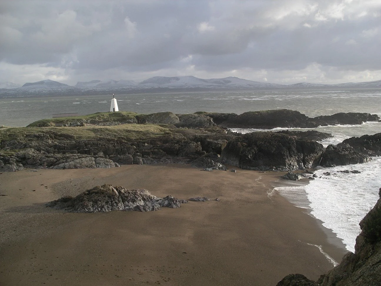 A white lighthouse tower on rocks above a sandy cove, with mountains beyond