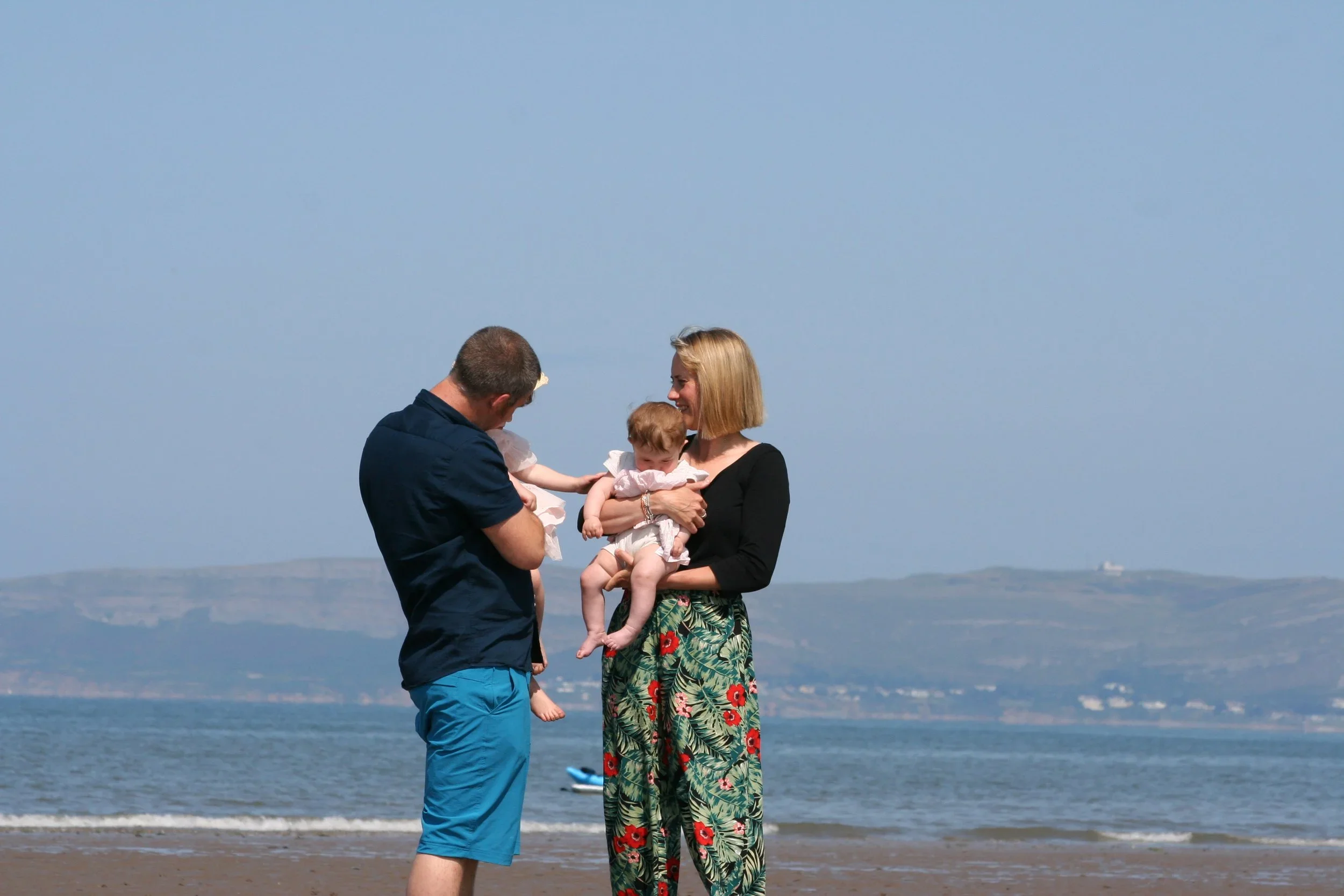 A couple holding their baby on a sunny beach, with hills across the bay