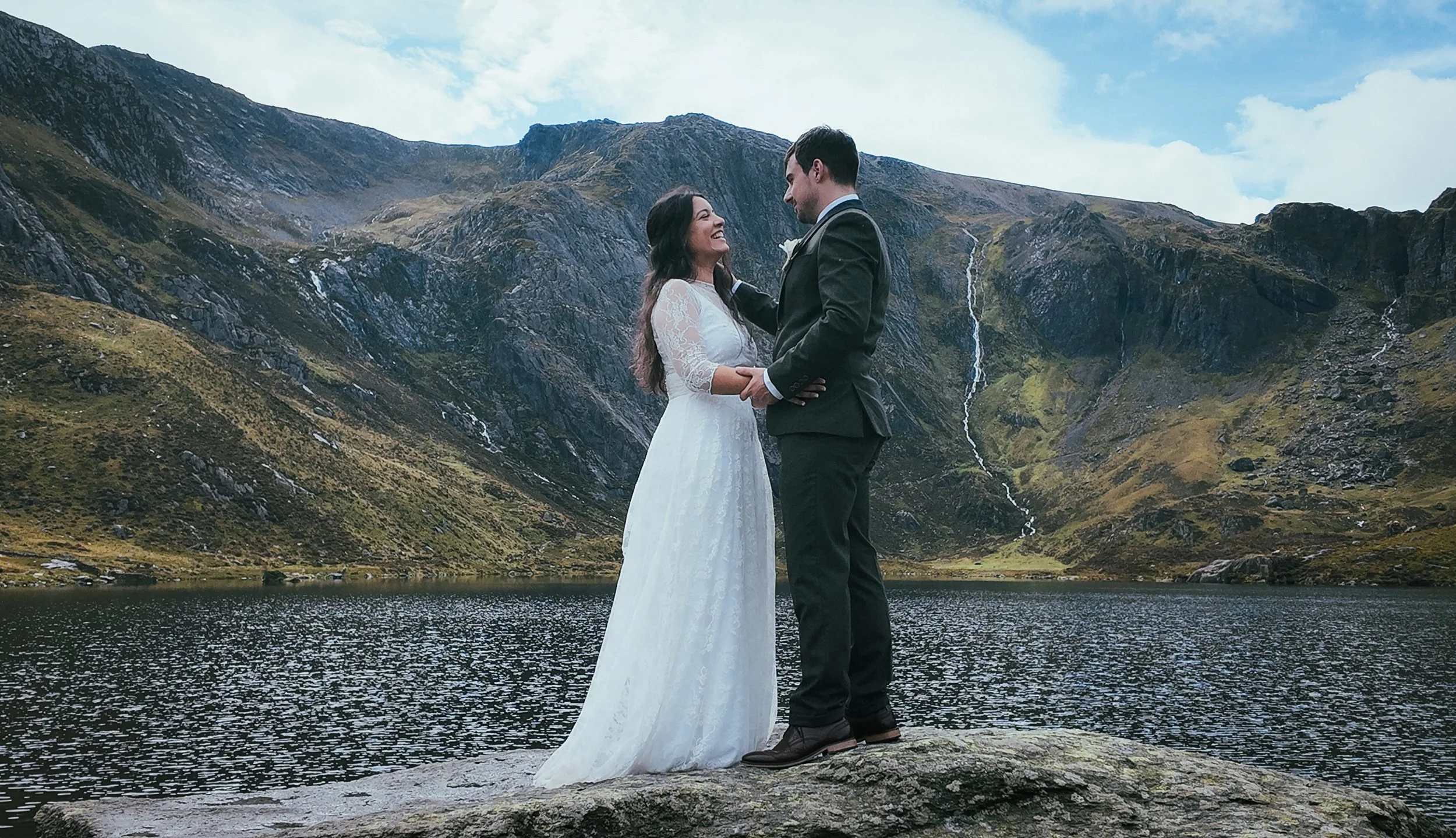 A couple smiling at each other on a rock beside a mountain lake