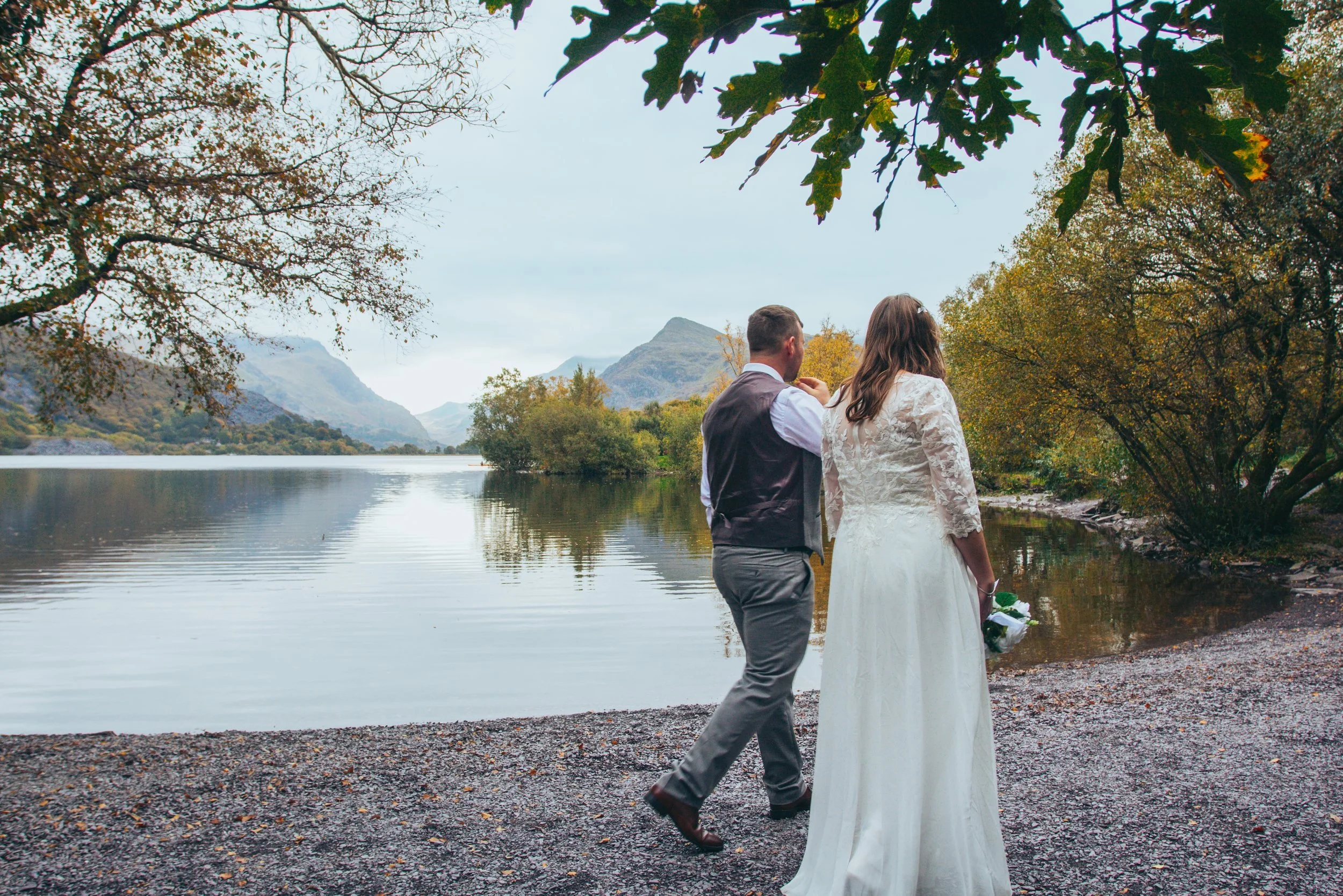 A couple walking along a lake shore beneath trees