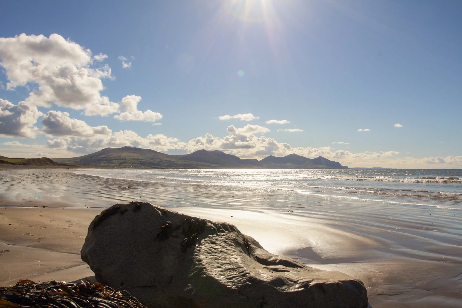 Sunlight over a sandy beach at low tide, with a large rock in the foreground and hills across the bay