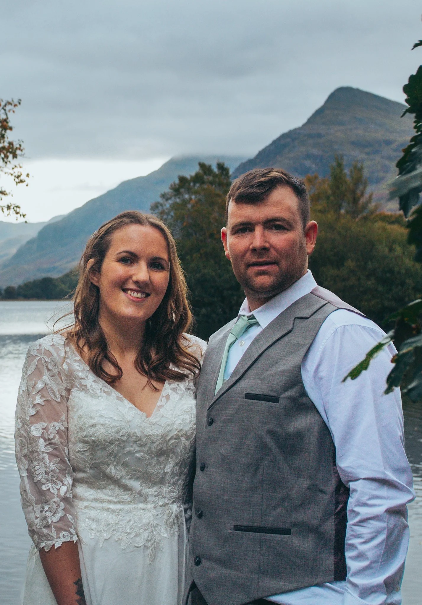 A couple smiling together beside a lake with mountains behind them