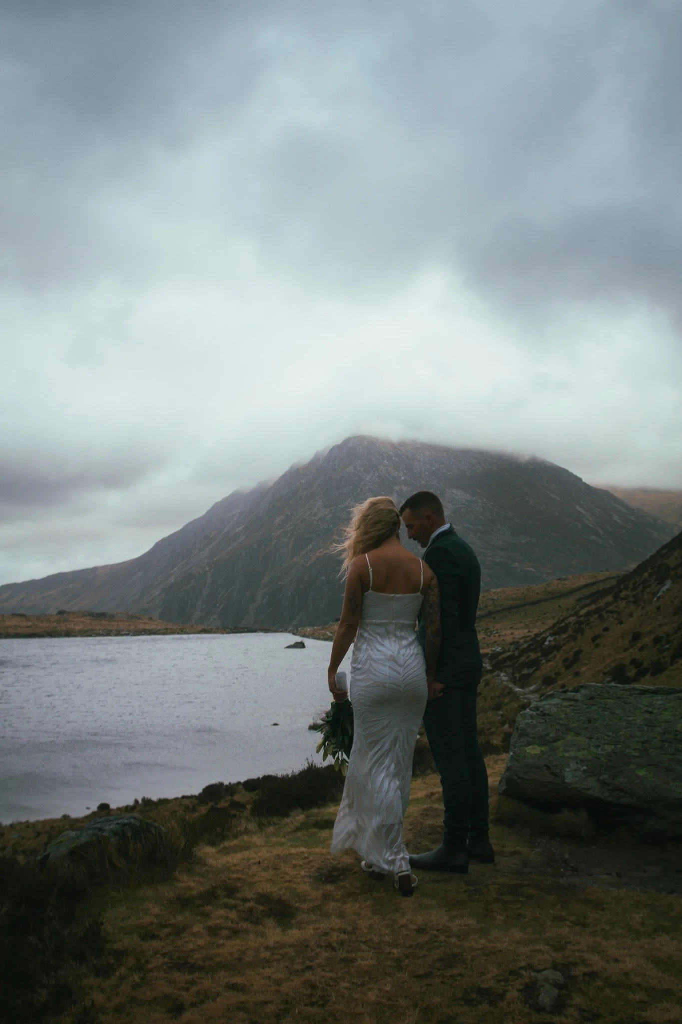 Couple standing on a lakeside ridge beneath a dark mountain