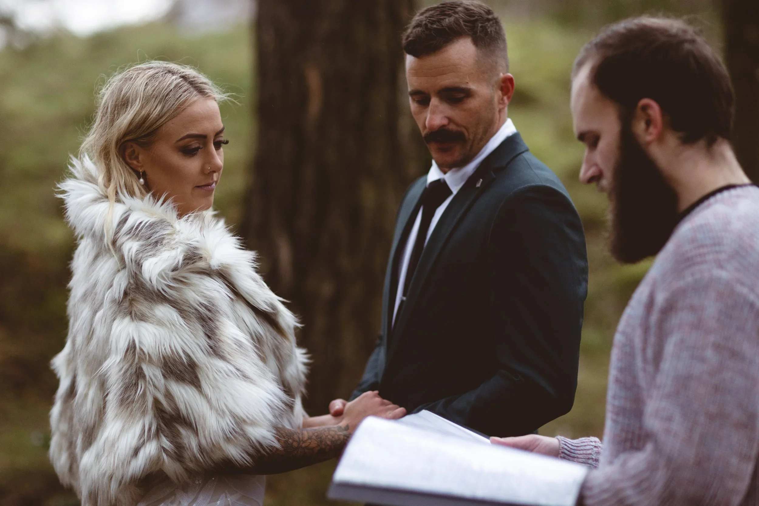 A bride in a fur wrap and her groom holding hands during a woodland elopement ceremony, with a celebrant beside them