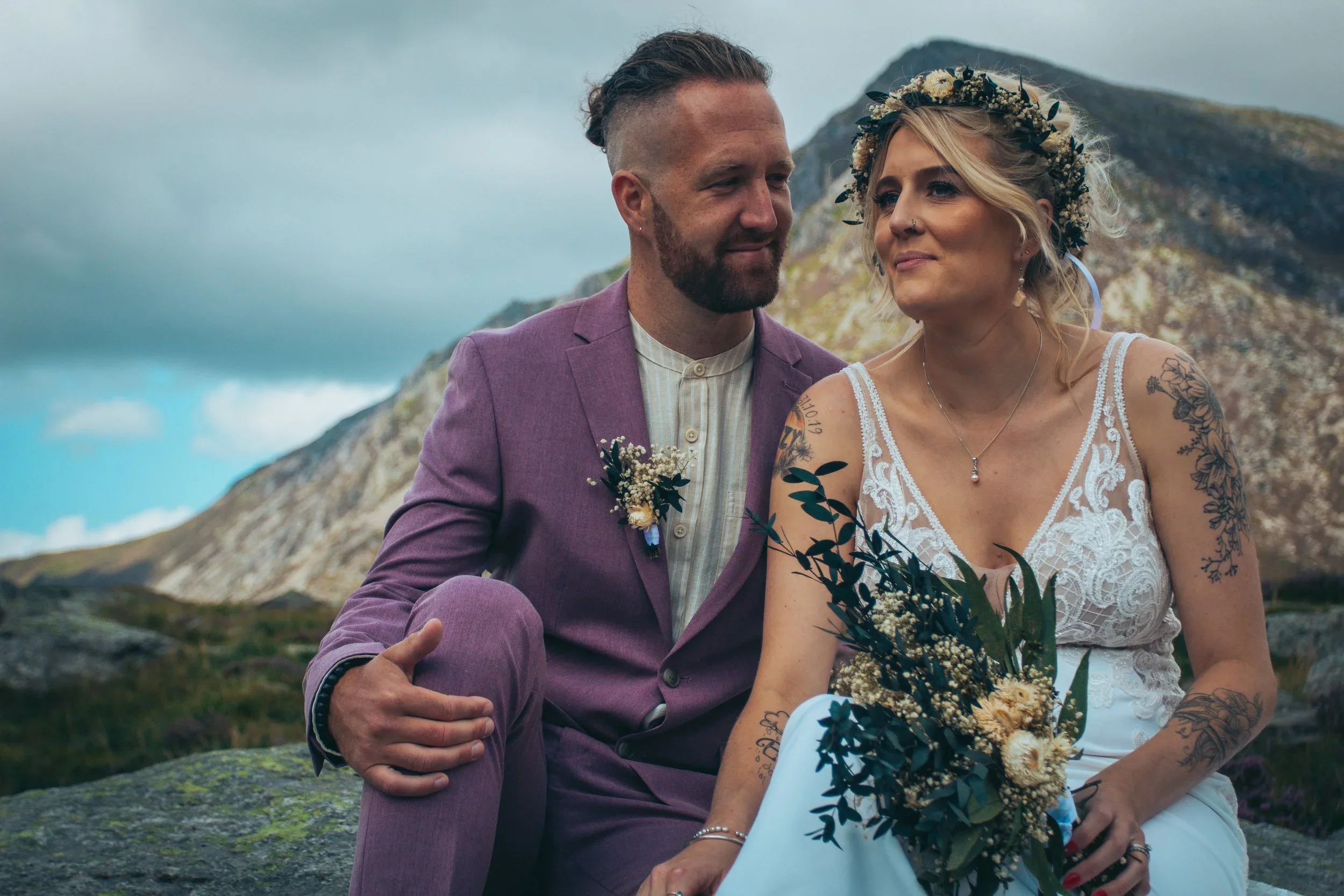 A smiling groom in a lilac suit and a bride in a flower crown holding her bouquet, with mountains behind them