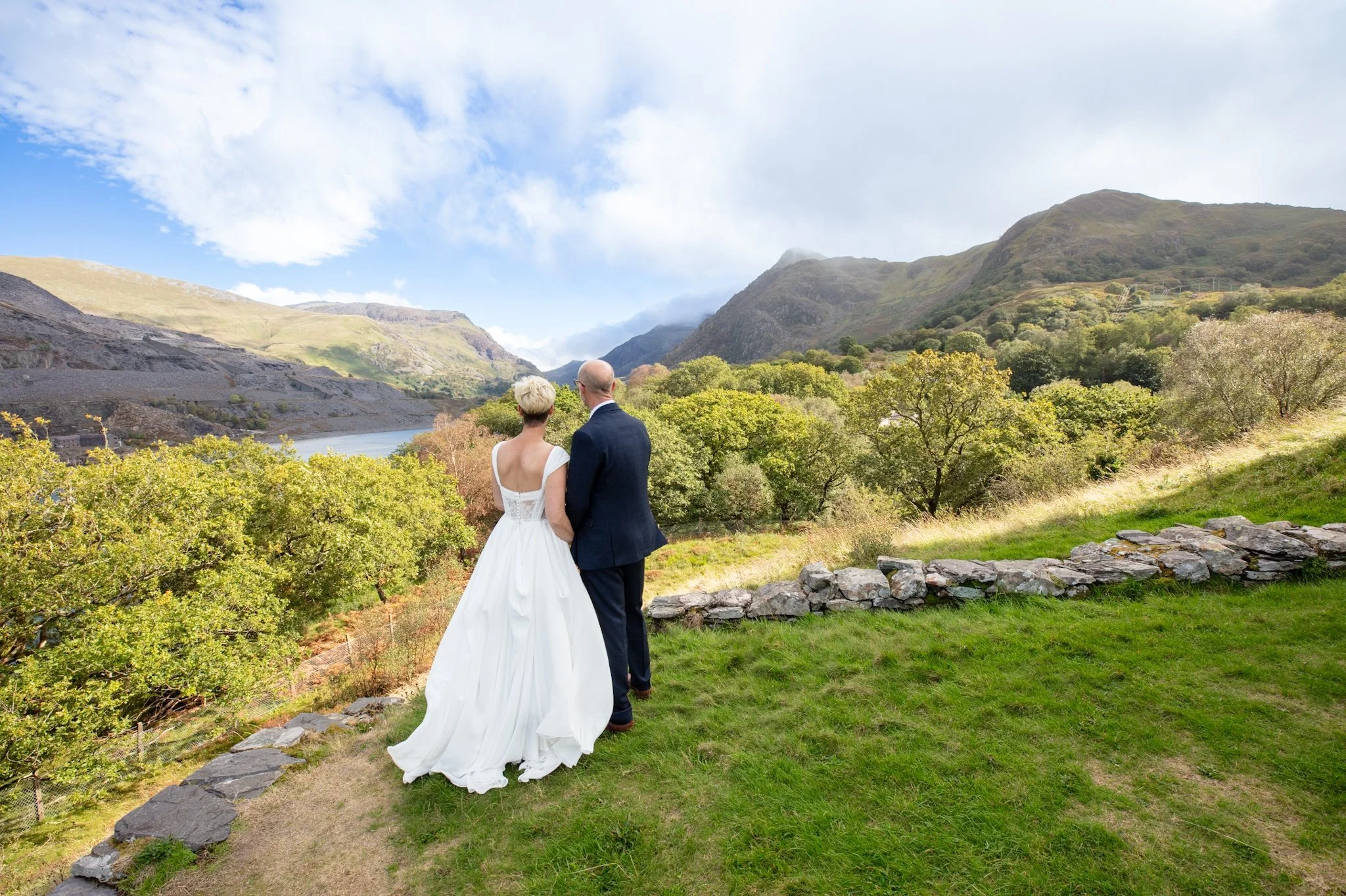 A couple seen from behind on a grassy hillside overlooking a lake and mountains