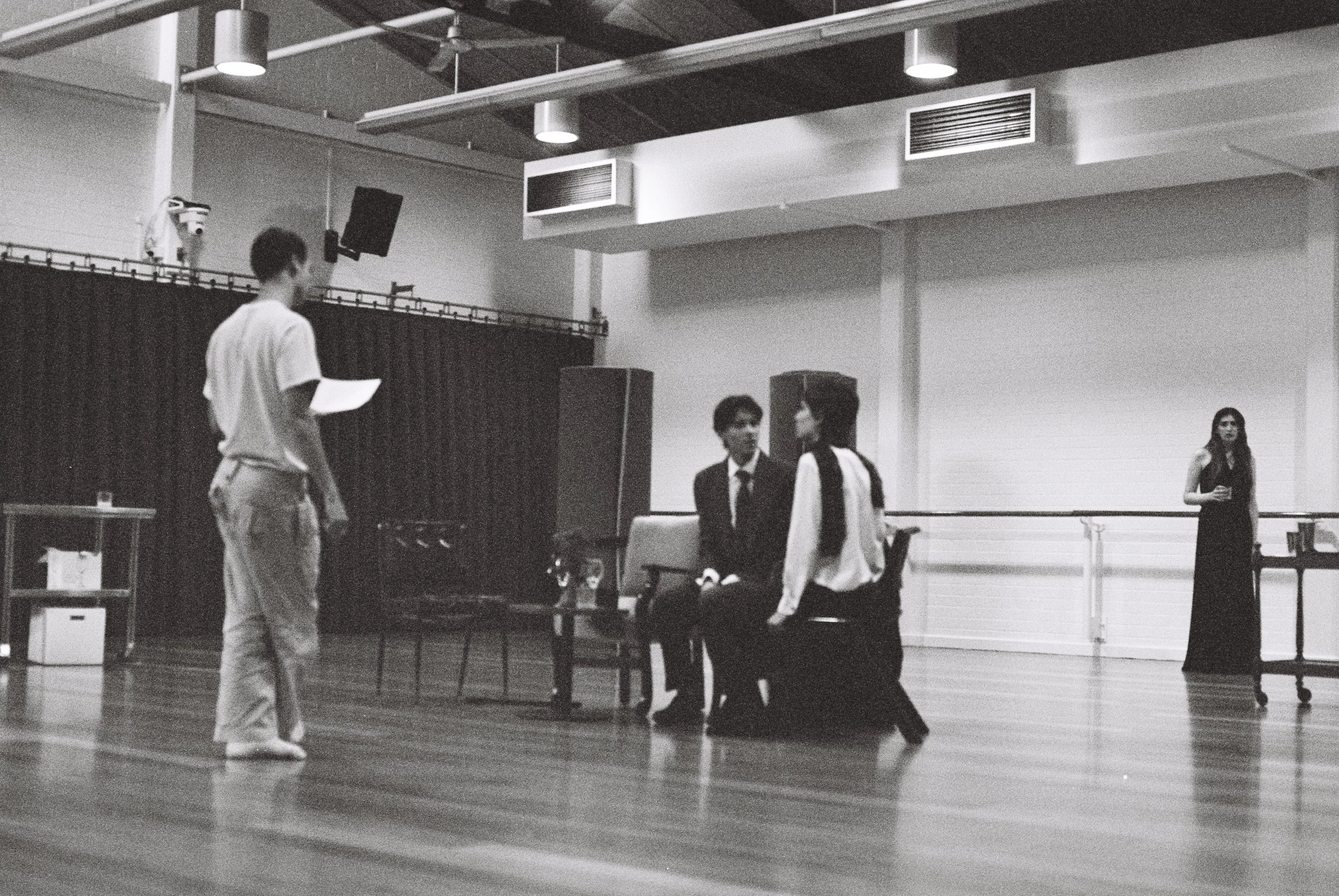 A black and white photo shows a dance studio with wooden floors and large windows. Several people are present, with a group of three sitting and talking, and a person standing nearby. In the background, a mirror and objects are visible, indicating a dance or rehearsal setting.