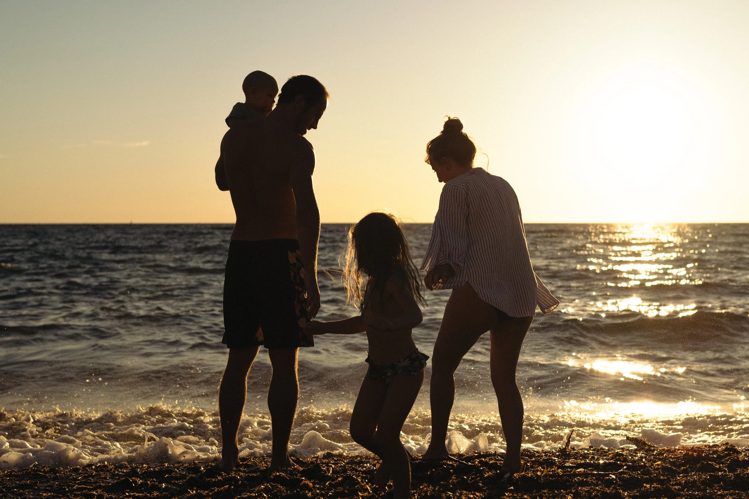Une famille au bord de la mer au coucher du soleil, comprenant deux adultes et deux enfants, en silhouette contre le ciel brillant par le soleil couchant.