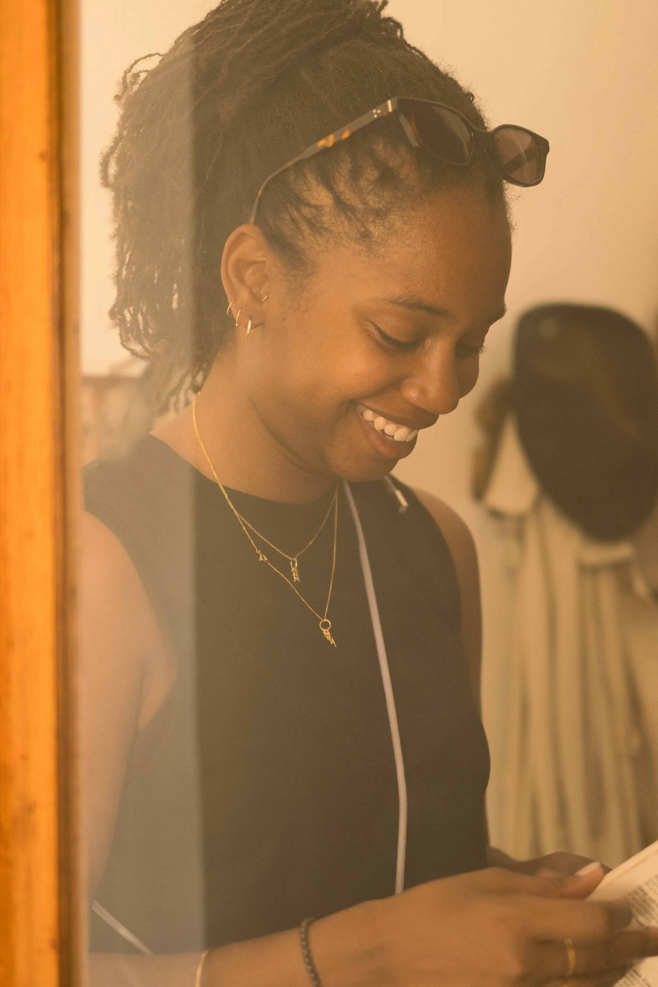 Jeune femme souriante portant des lunettes de soleil sur la tête, regardant un livre, avec des boucles d'oreilles dorées, un collier et un bracelet, à l'intérieur.