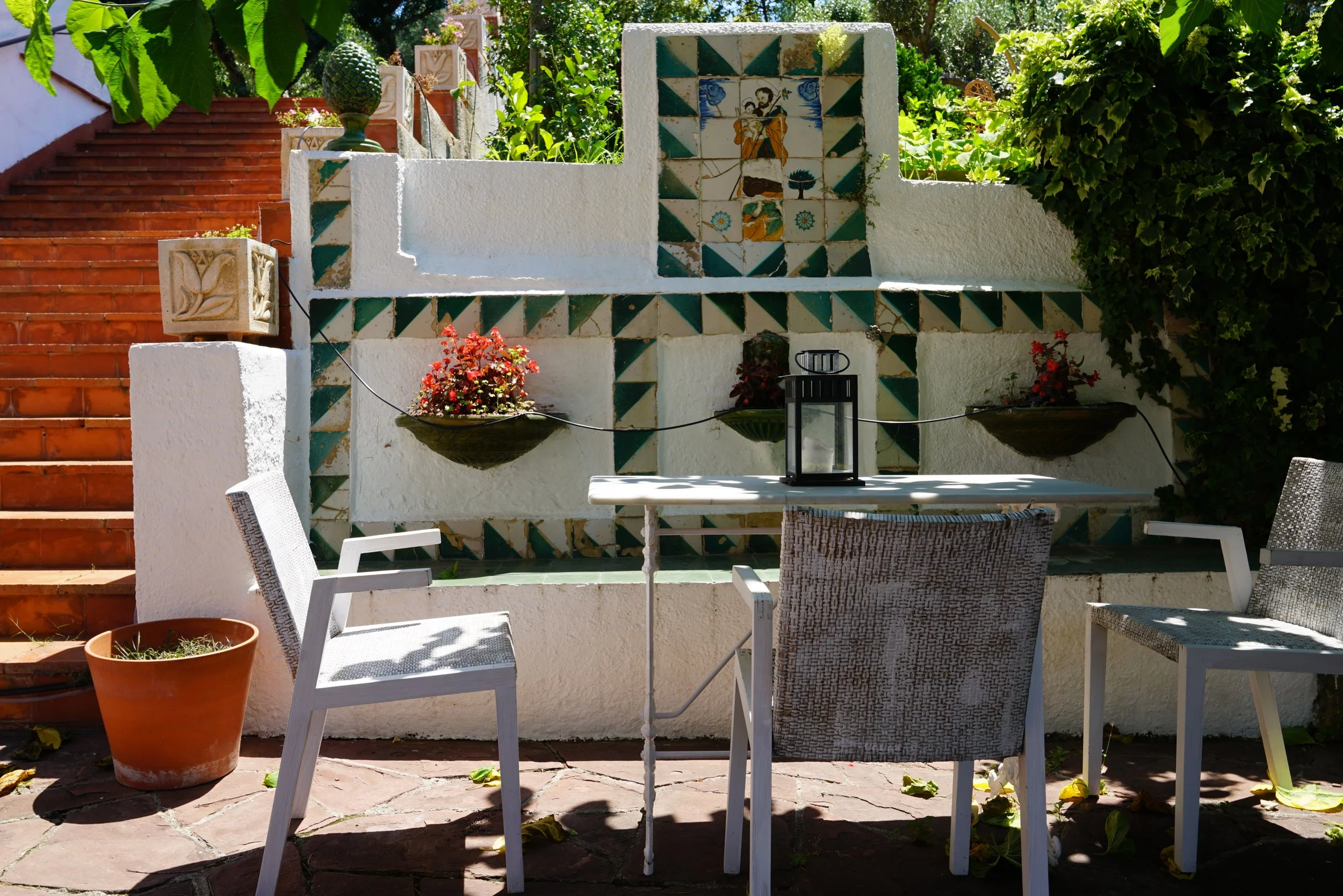 Outdoor patio with a white table and chairs, decorative tiles on the wall, potted plants, green shutters, and a staircase with red tiles in the background, surrounded by greenery.