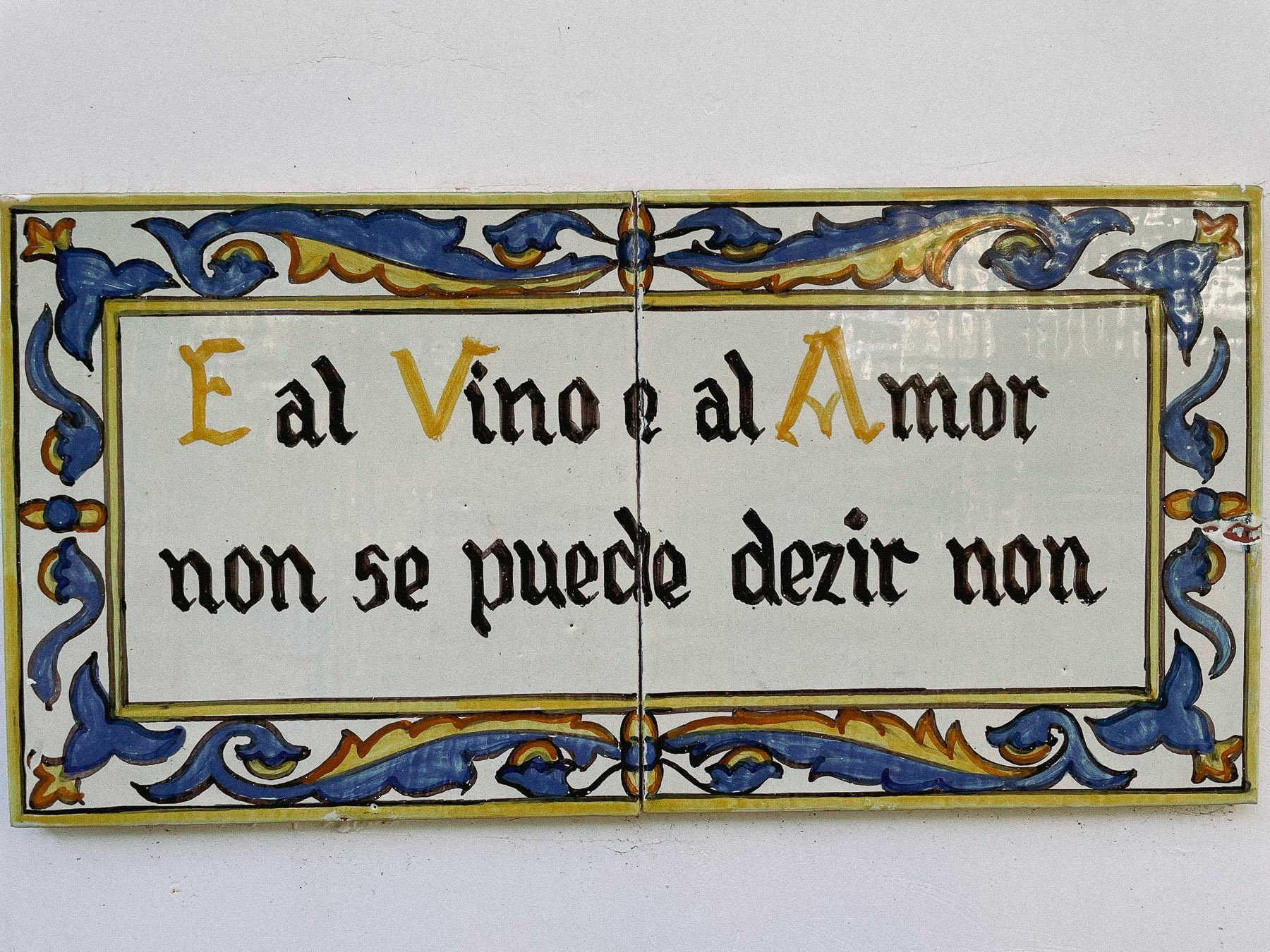 Decorative tile with a border of blue and yellow scrollwork surrounding a quote in Spanish that reads 'El vino al Amor non se puede dezir non'.