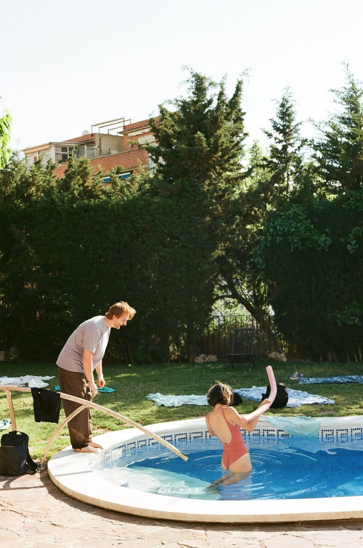 A person with short hair and a swimsuit is in a backyard swimming pool, holding a pink pool noodle. A person with short hair, wearing a white t-shirt and brown shorts, is standing at the pool's edge, leaning over slightly. There are towels and clothes laid out on the grass, surrounded by trees and a fence, with a house visible in the background.