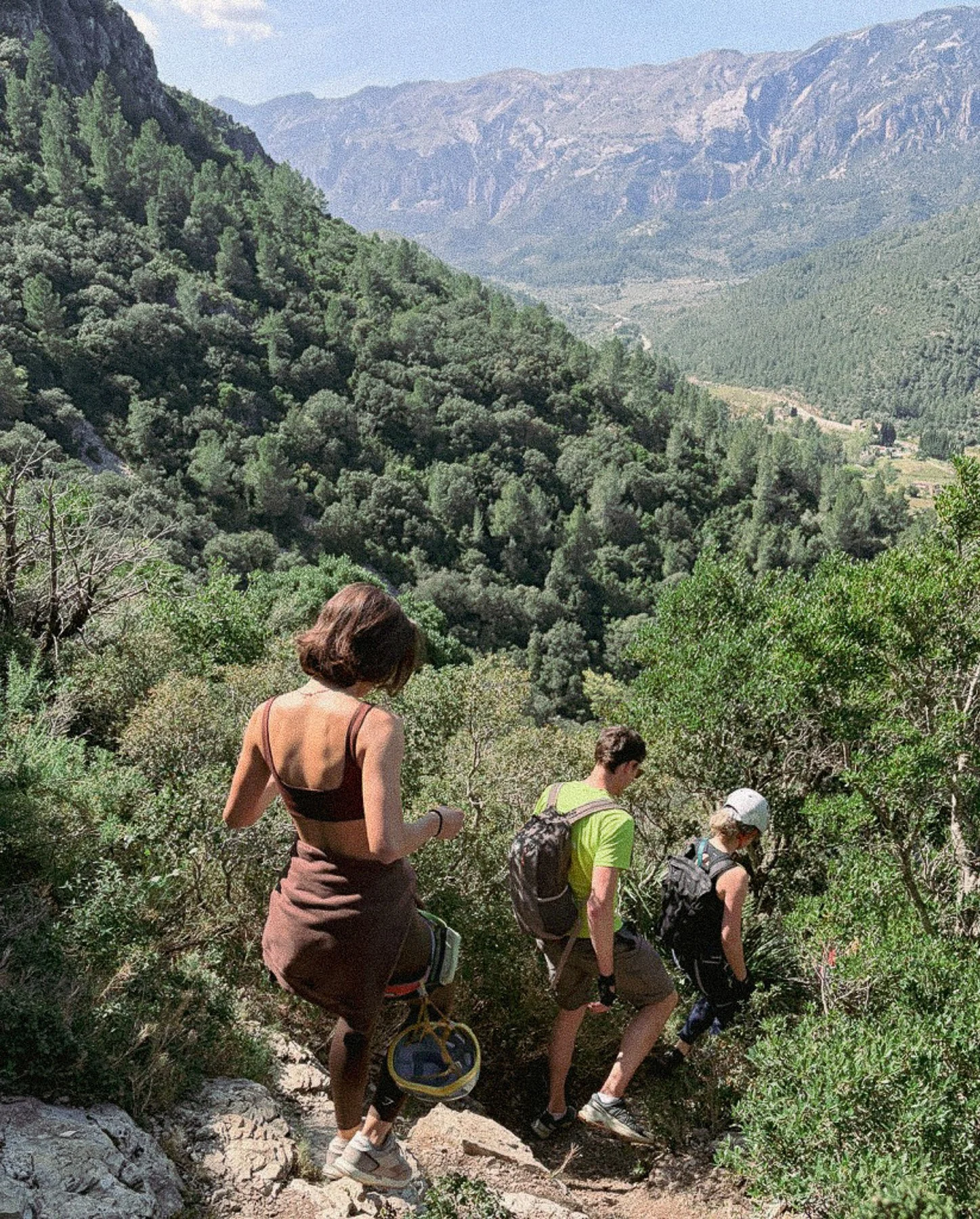 Group of four hikers walking along a trail in a mountain valley surrounded by green trees and distant mountains.