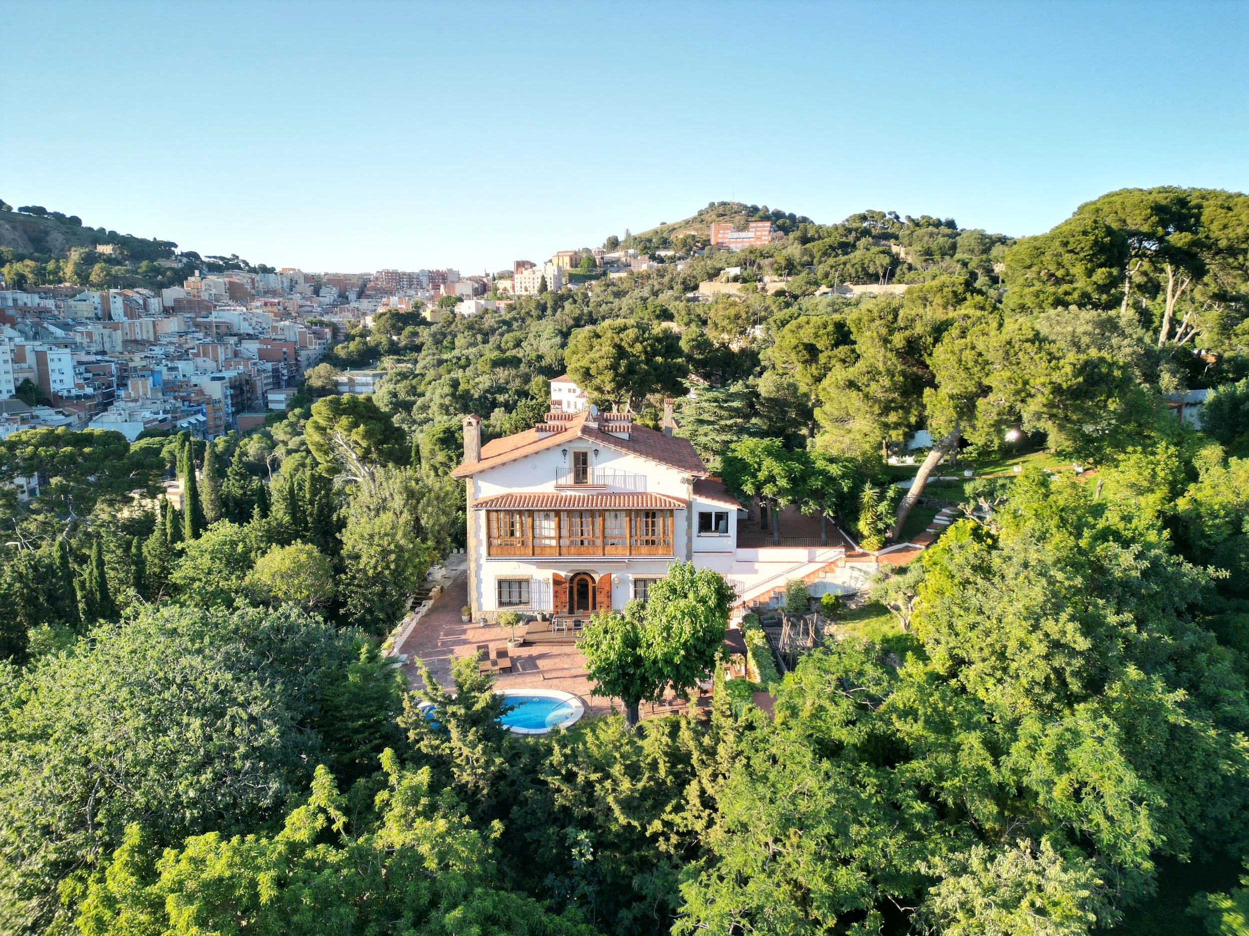 A landscape view of a city built on hills, with numerous trees and a house with a red-tiled roof in the foreground, and a swimming pool visible beside it.