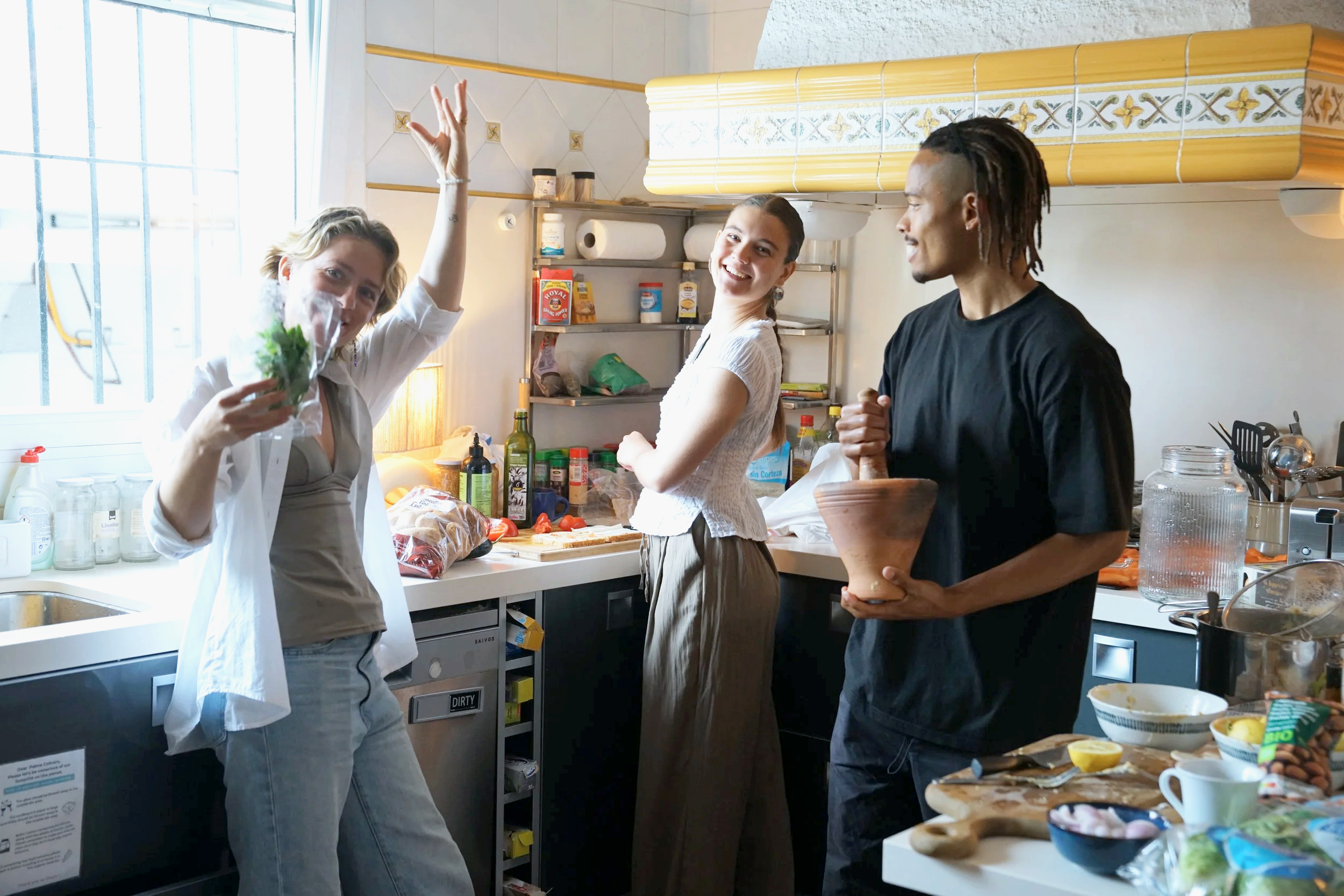 Colivers are in a kitchen preparing food for community dinners, smiling and engaging with each other; the woman on the left is holding herbs, the woman in the middle is smiling with arms crossed, and the man on the right is holding a bowl.