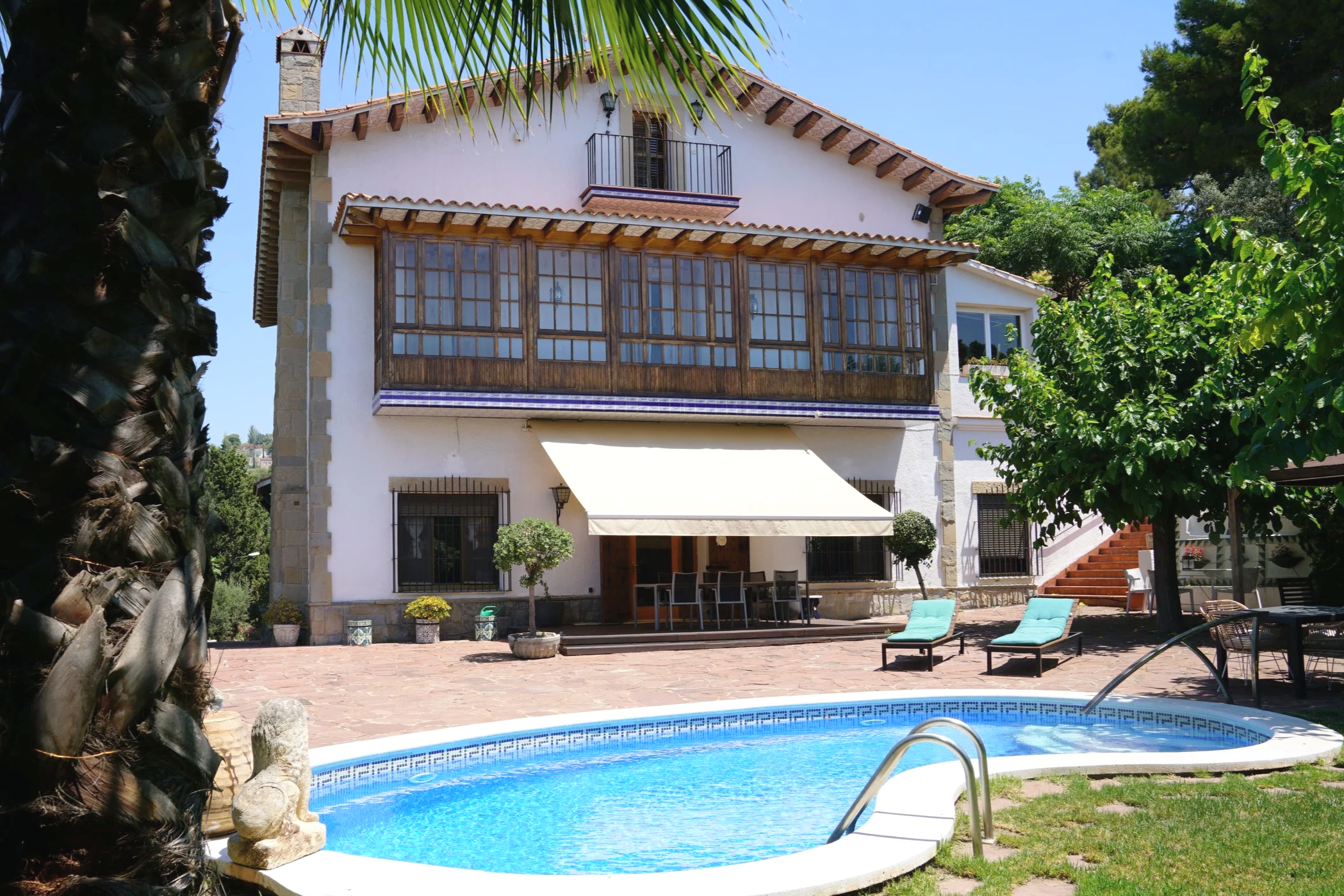 Luxury house in Barcelona for coliving with a large garden swimming pool, lounge chairs, and outdoor seating area, surrounded by lush greenery and trees.