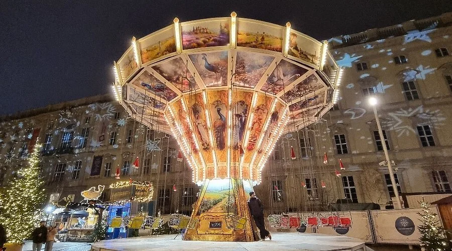 A lit amusement park swing ride at night with colorful artwork on the canopy, surrounded by holiday decorations and small Christmas trees.