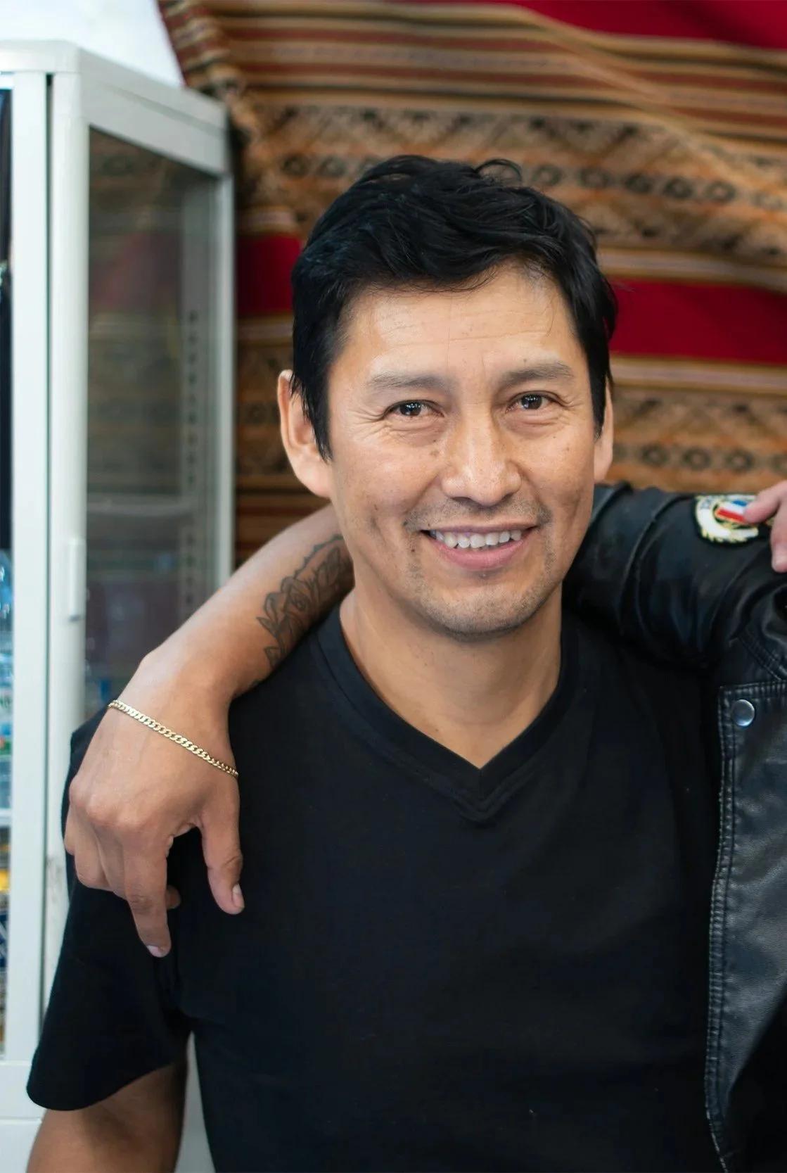 A smiling man with black hair and a light beard, wearing a black shirt, with someone else's arm around his shoulder, in front of a colorful patterned background.