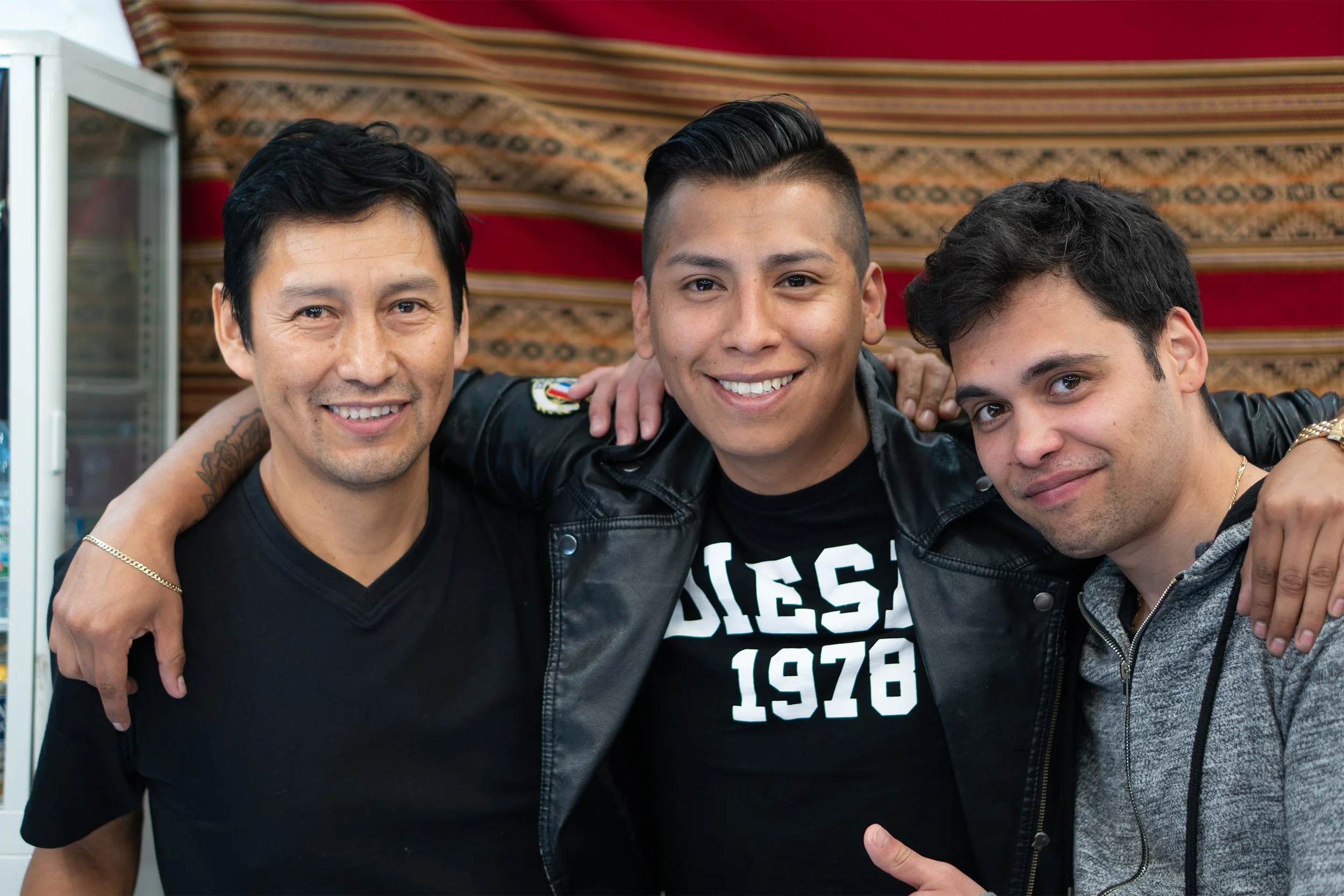 Three men smiling with arms around each other, wearing black shirts and jackets, standing in front of a decorated background.