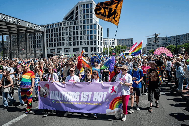 Crowd of people participating in a pride parade, holding rainbow flags, banners, and wearing colorful clothing and accessories in Berlin.