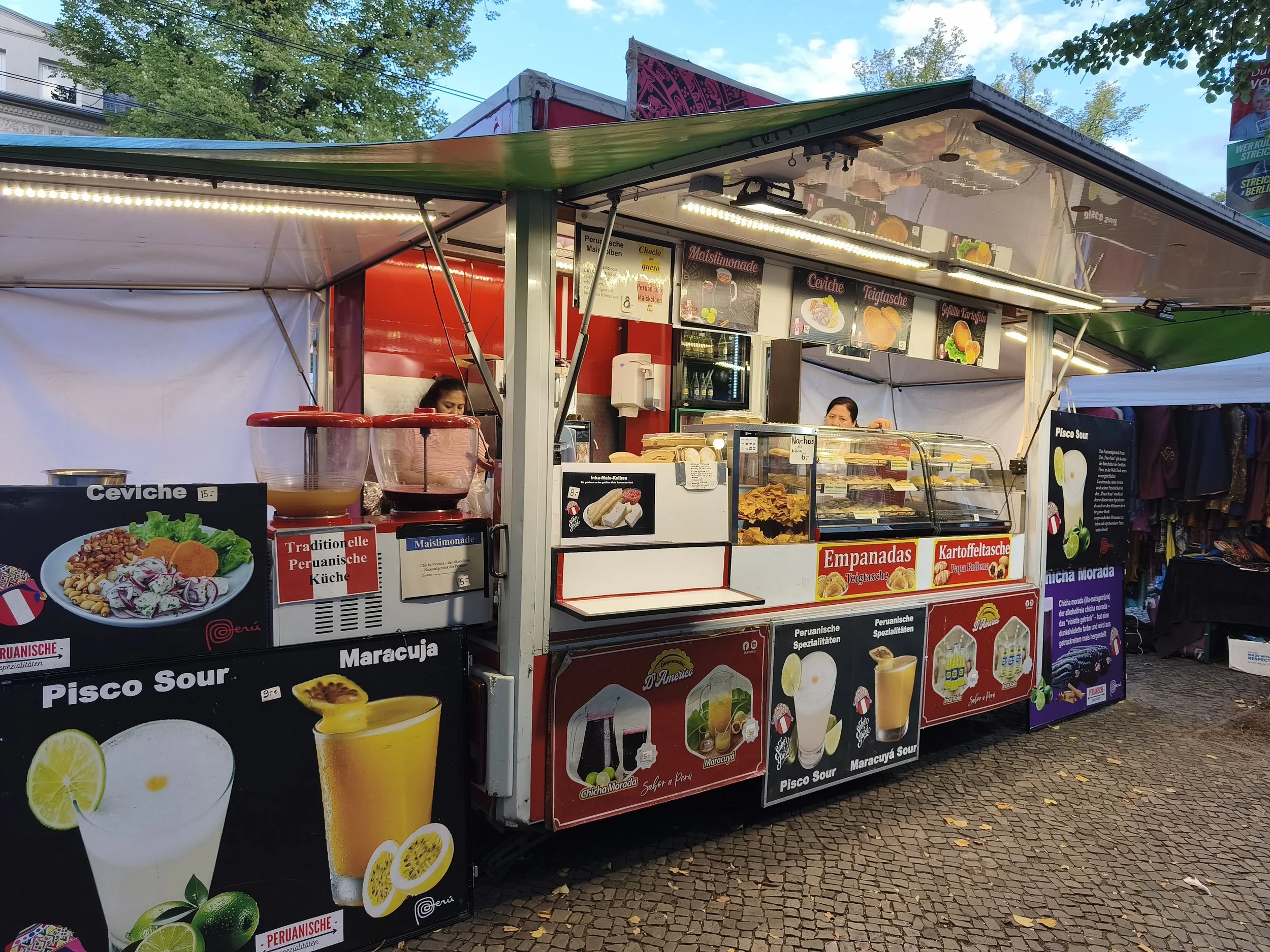Food stall selling Peruvian dishes, with signs displaying ceviche, empanadas, and passion fruit drinks, set outdoors with trees and a blue sky in the background.