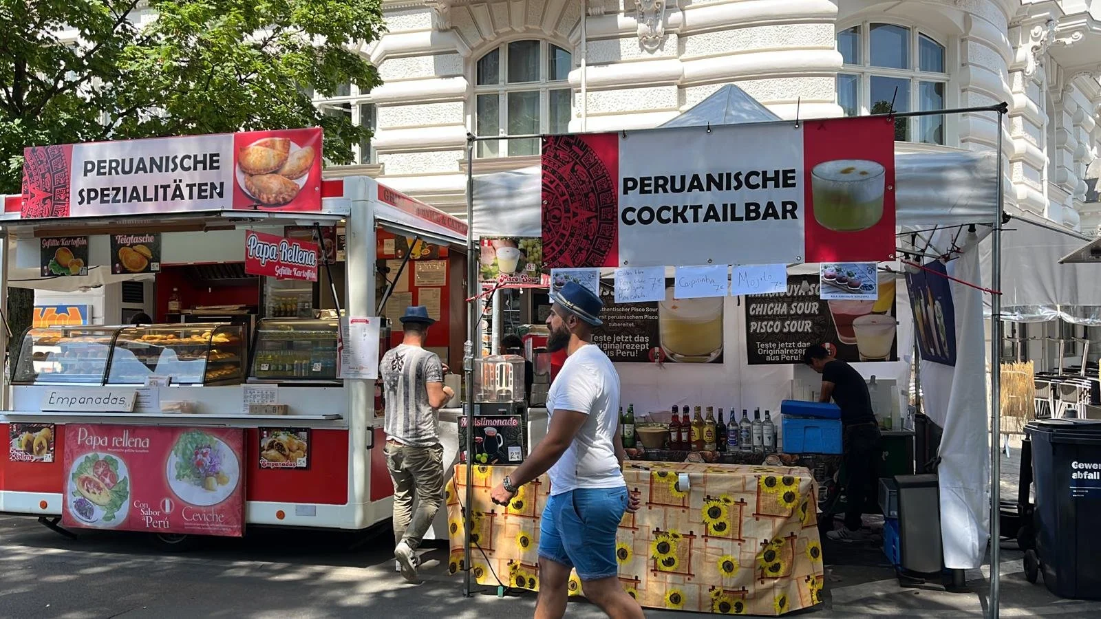 Peruvian street food stall selling empanadas and drinks at an outdoor market in front of a historic building, with signs displaying Peruvian specialties, cocktails, and colorful menus.