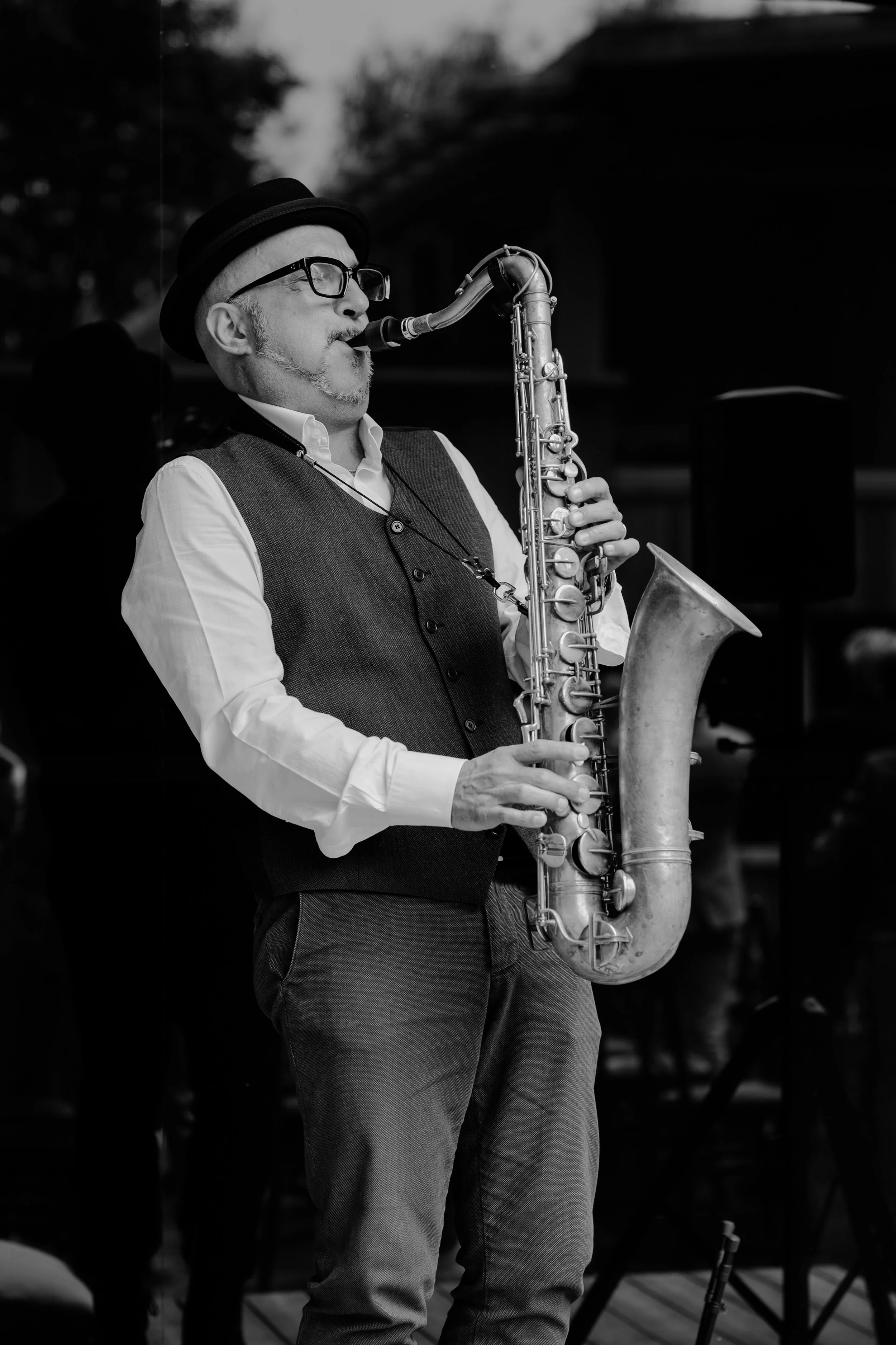 A black and white photo of a man playing a saxophone. He is wearing glasses, a hat, a vest over a white shirt, and pants. The background is dark and studio-like.