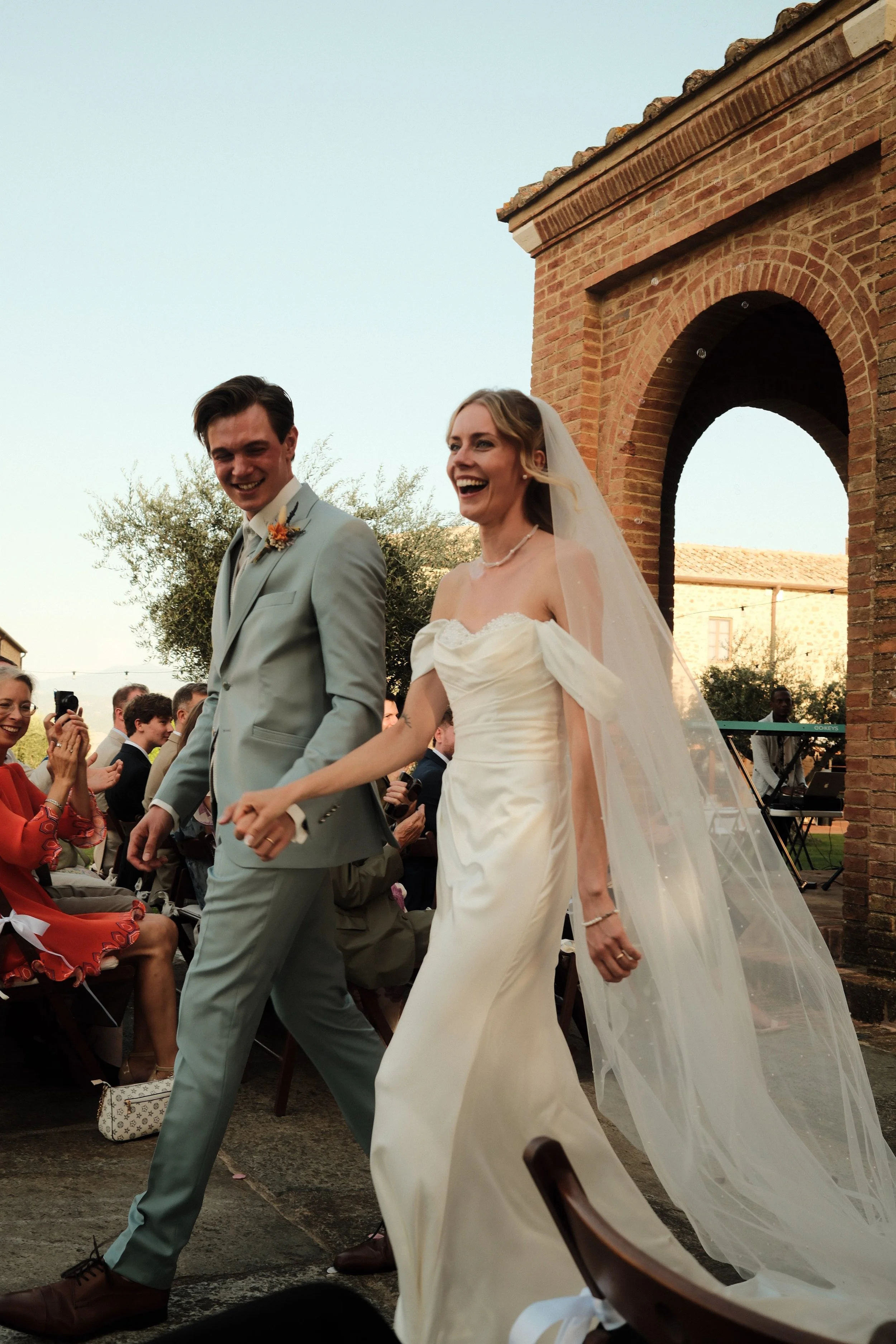 Lachend bruidspaar loopt hand in hand na de buitenceremonie bij een stenen kapel in Toscane.