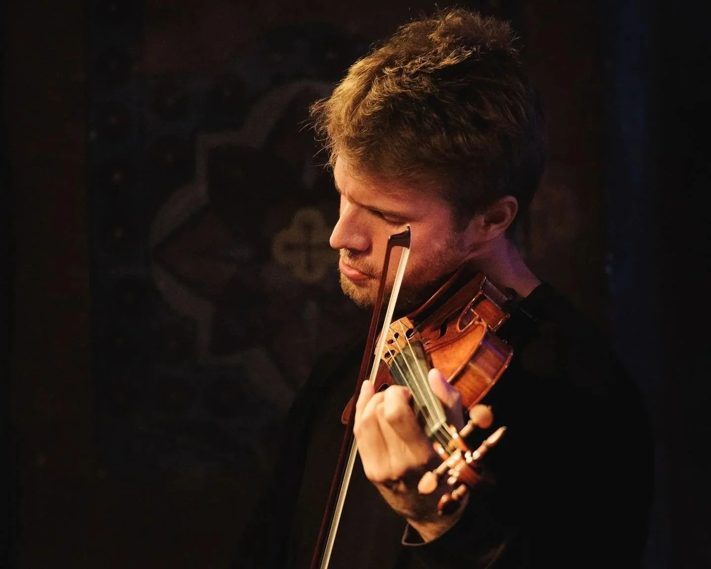 Jeune homme jouant du violon, yeux fermés, dans un lieu sombre.
