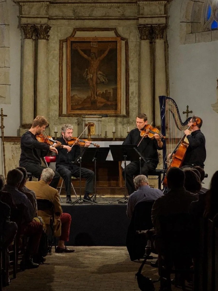 Quatre musiciens jouant du violon, de l'alto, du violoncelle et de la harpe lors d'un concert dans une église, avec un tableau religieux en arrière-plan et un public assis devant eux.