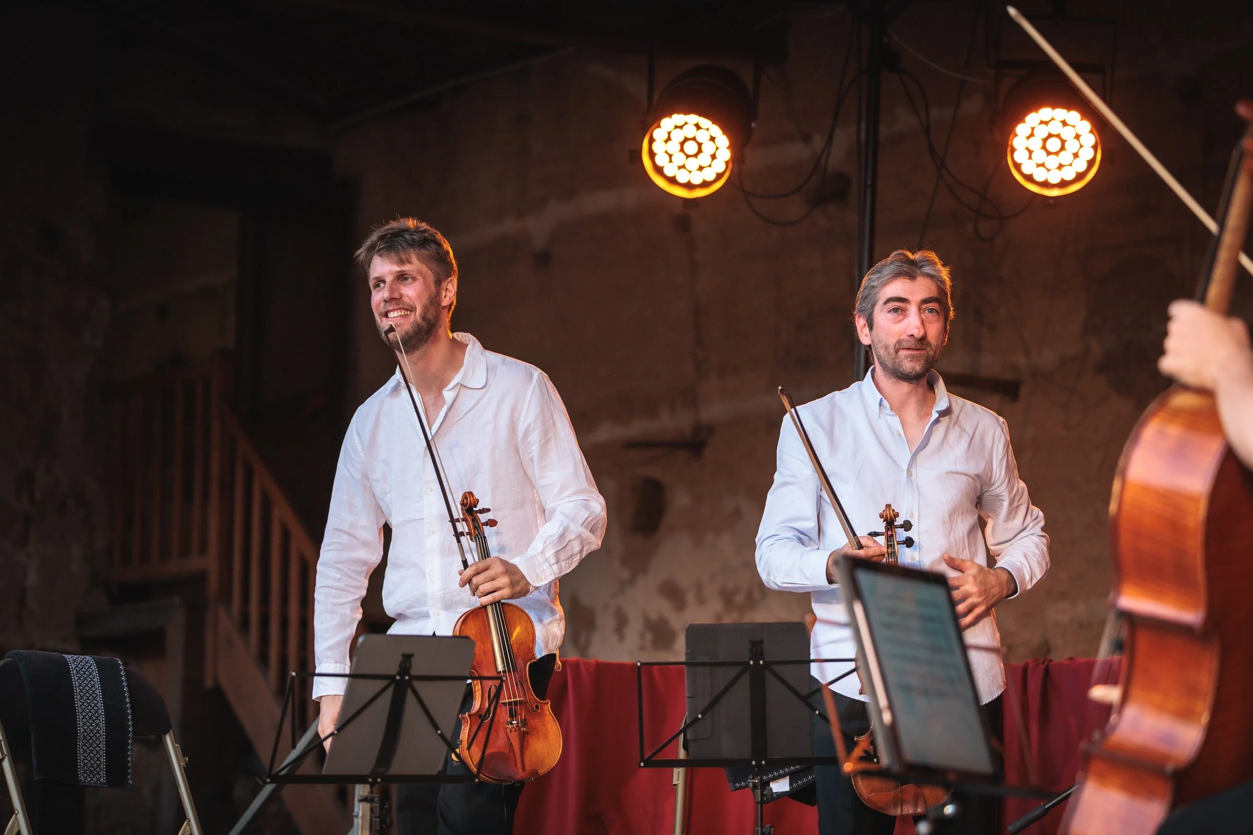 Deux hommes jouent du violon lors d'une performance musicale en intérieur, avec des lumières orange en arrière-plan.