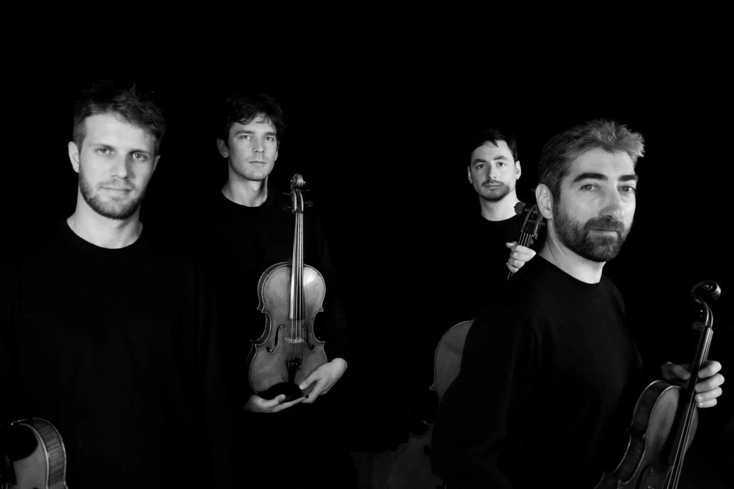 Quatre hommes tenant des violons, posant dans un fond noir, en noir et blanc.