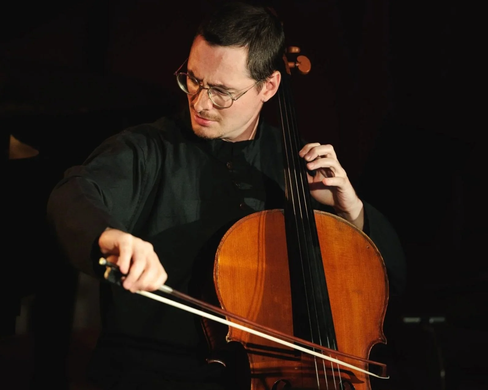 Un homme jouant du violoncelle, portant des lunettes et concentré, dans un environnement sombre.