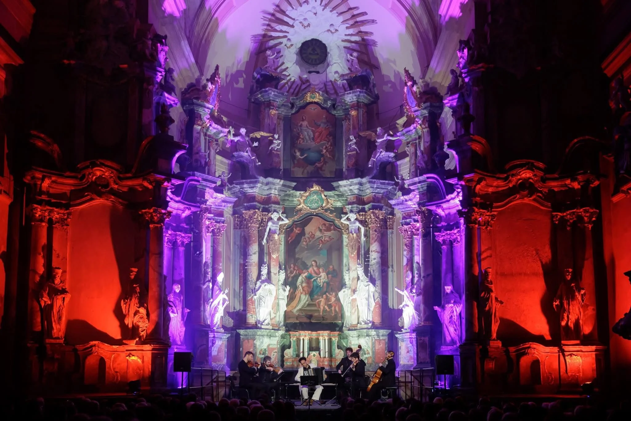 Une scène religieuse dans une église ornée de sculptures et de peintures baroques, avec un groupe de musiciens jouant devant un autel illuminé par des lumières violettes et rouges.