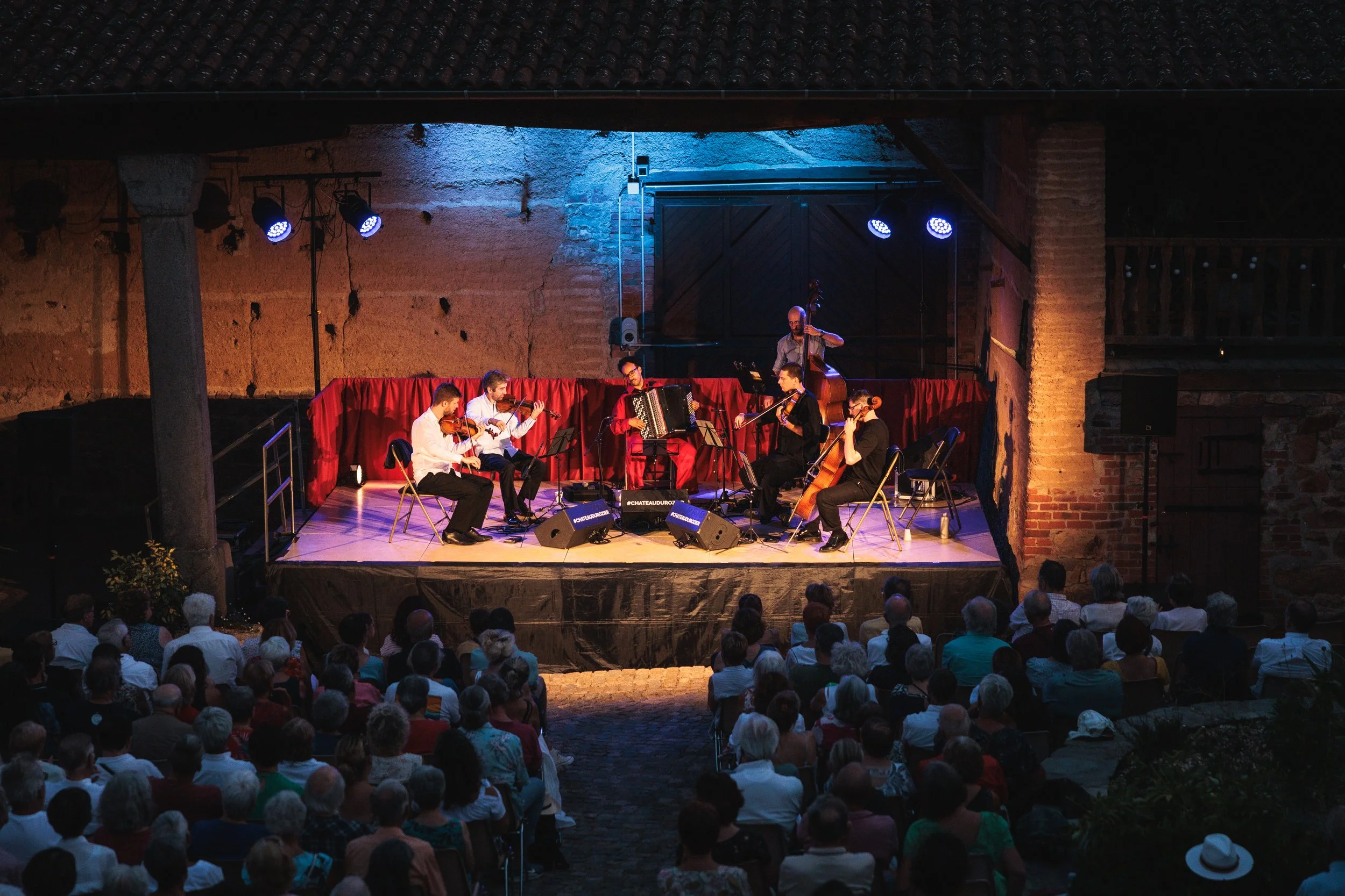 Un groupe de musiciens joue lors d'un concert en plein air, sous un grand toit en tuiles, devant une audience assise. Le groupe comprend des violonistes, un accordéoniste, un joueur de contrebasse et un violoncelliste, avec un fond de mur en pierre e