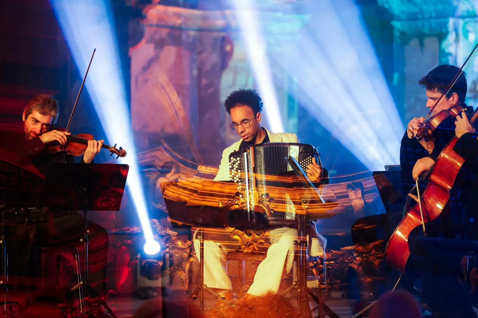 Trois musiciens jouent du violon et de l'accordéon sur scène, avec des lumières colorées en arrière-plan.