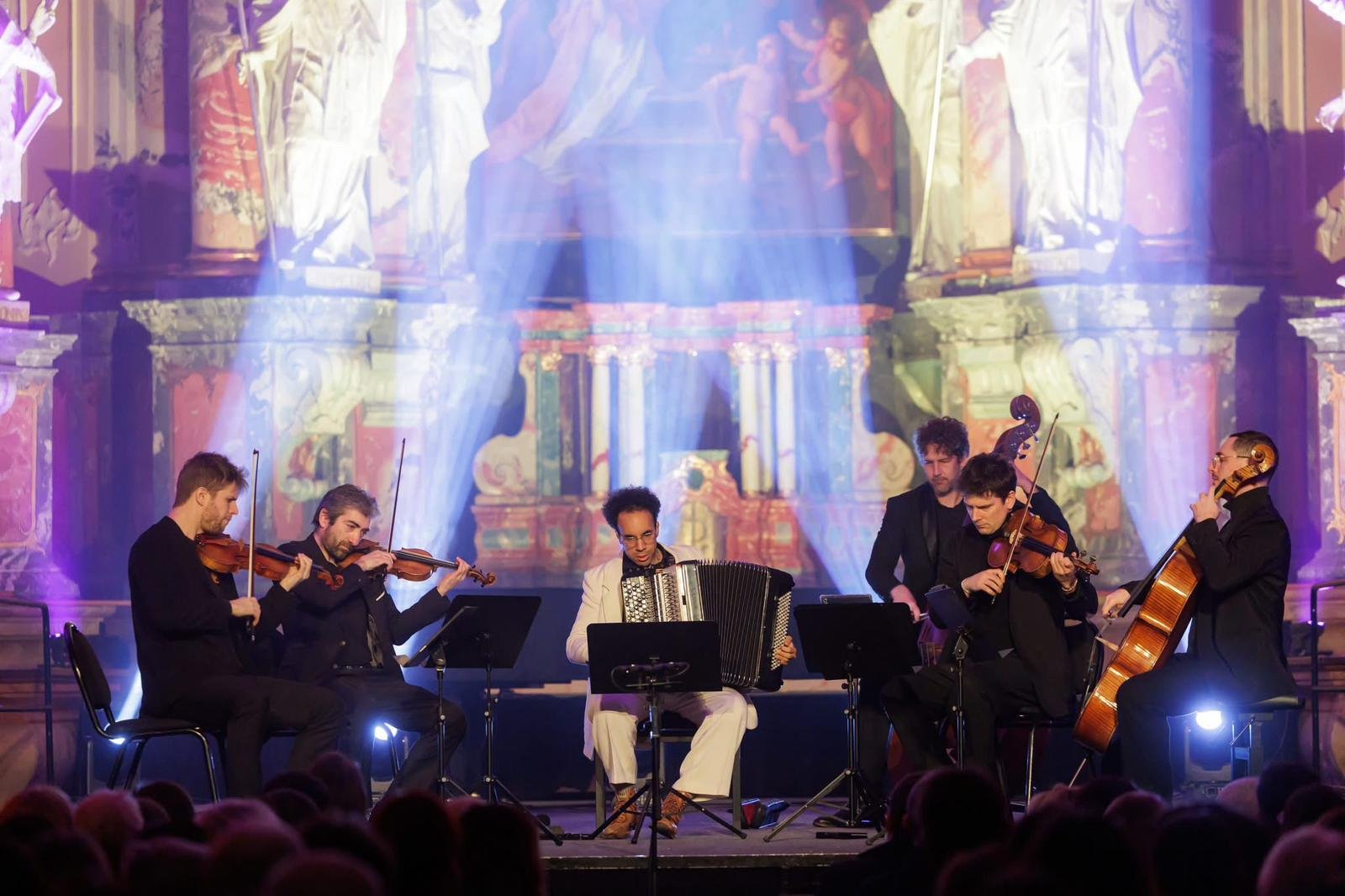 Groupe de musiciens jouant du violon, un joueur d'accordéon et un violoncelliste lors d'un concert dans une salle ornée de fresques et de colonnes.
