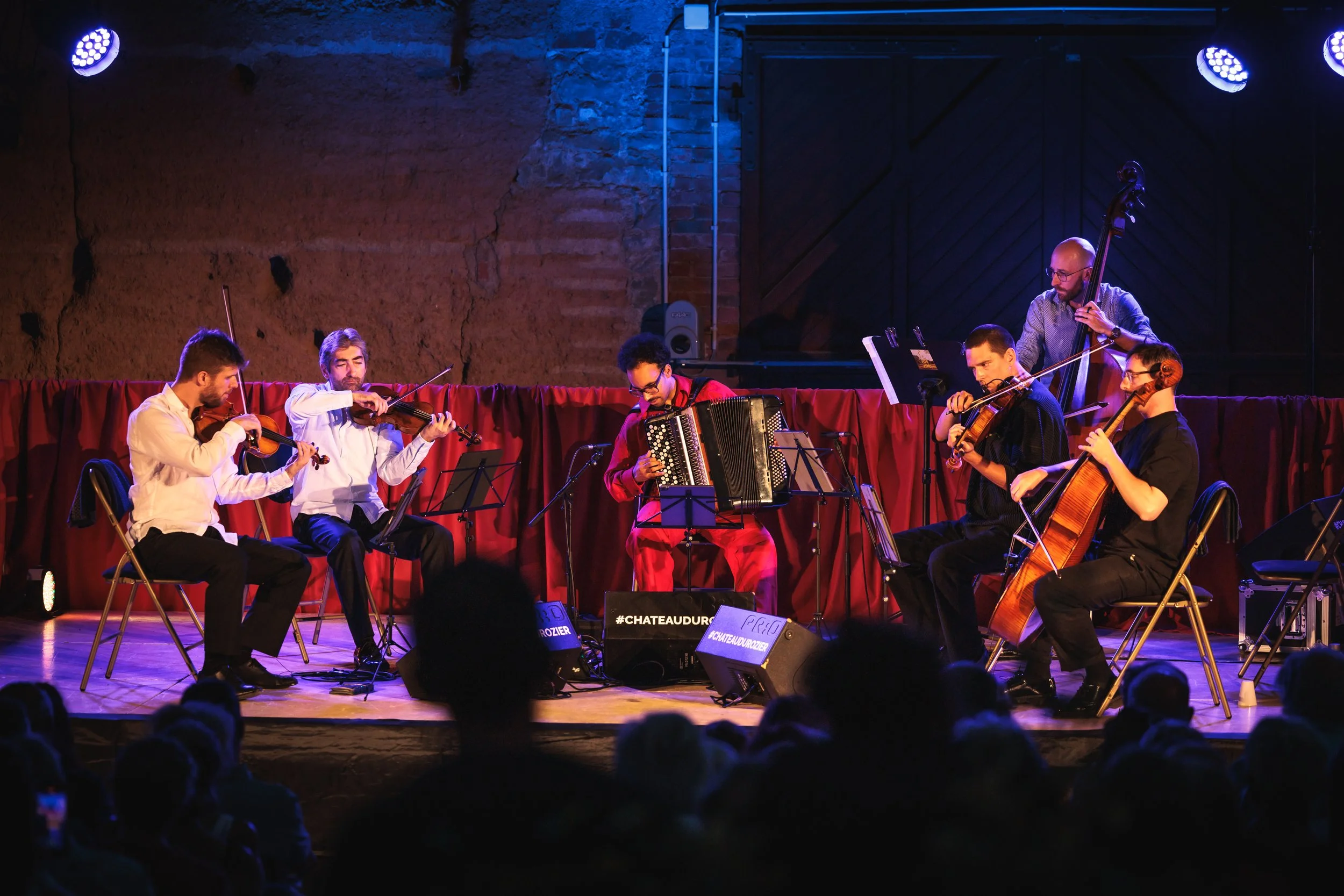 Un groupe de musiciens jouant sur scène, comprenant des violonistes, un violoncelliste et un accordéoniste, lors d'un concert avec éclairage bleu et rouge, dans un lieu avec murs en pierre et une grande porte en bois.