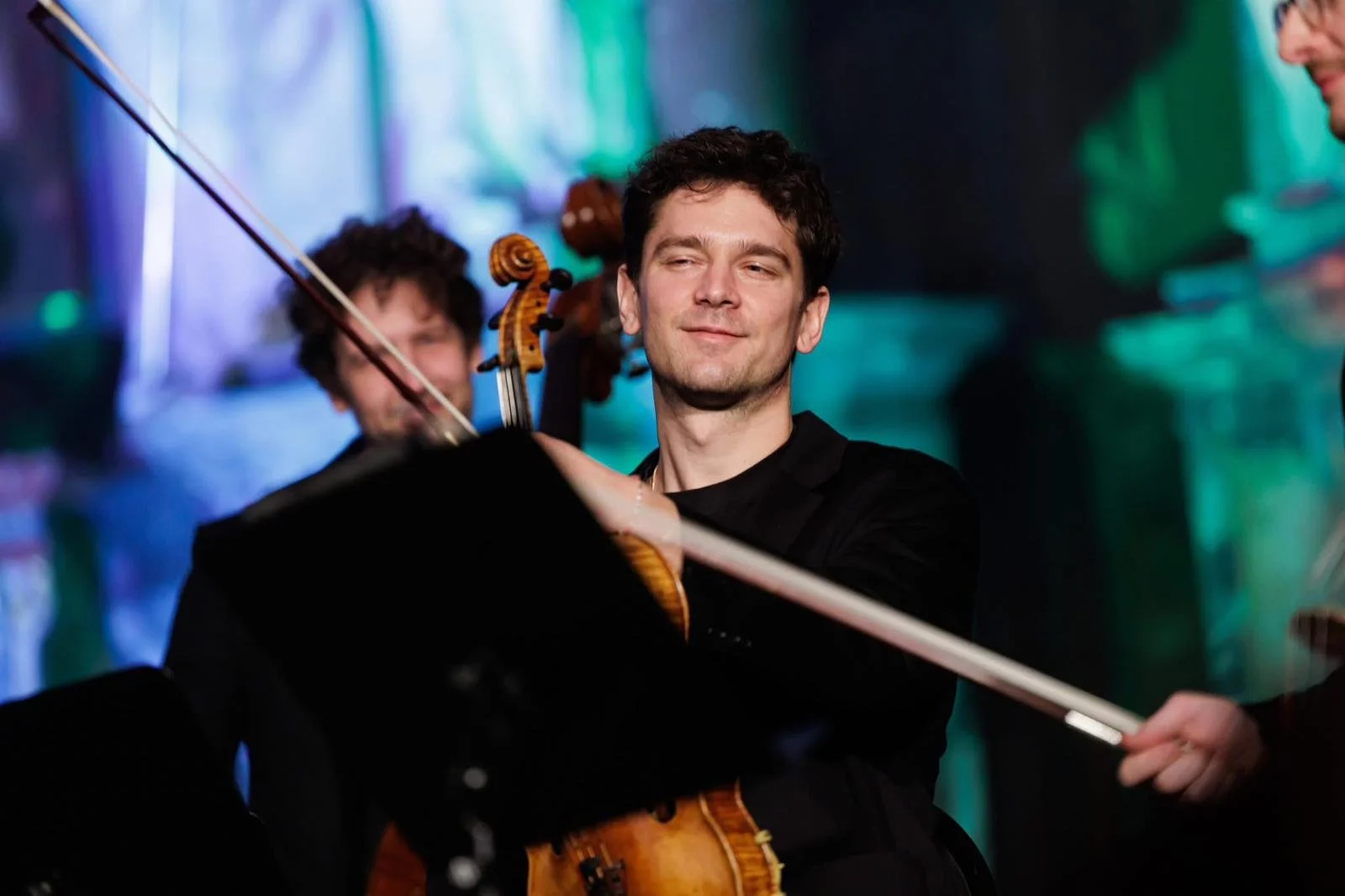 Musicien jouant du violon lors d'une représentation, avec un fond coloré et des autres musiciens en arrière-plan.