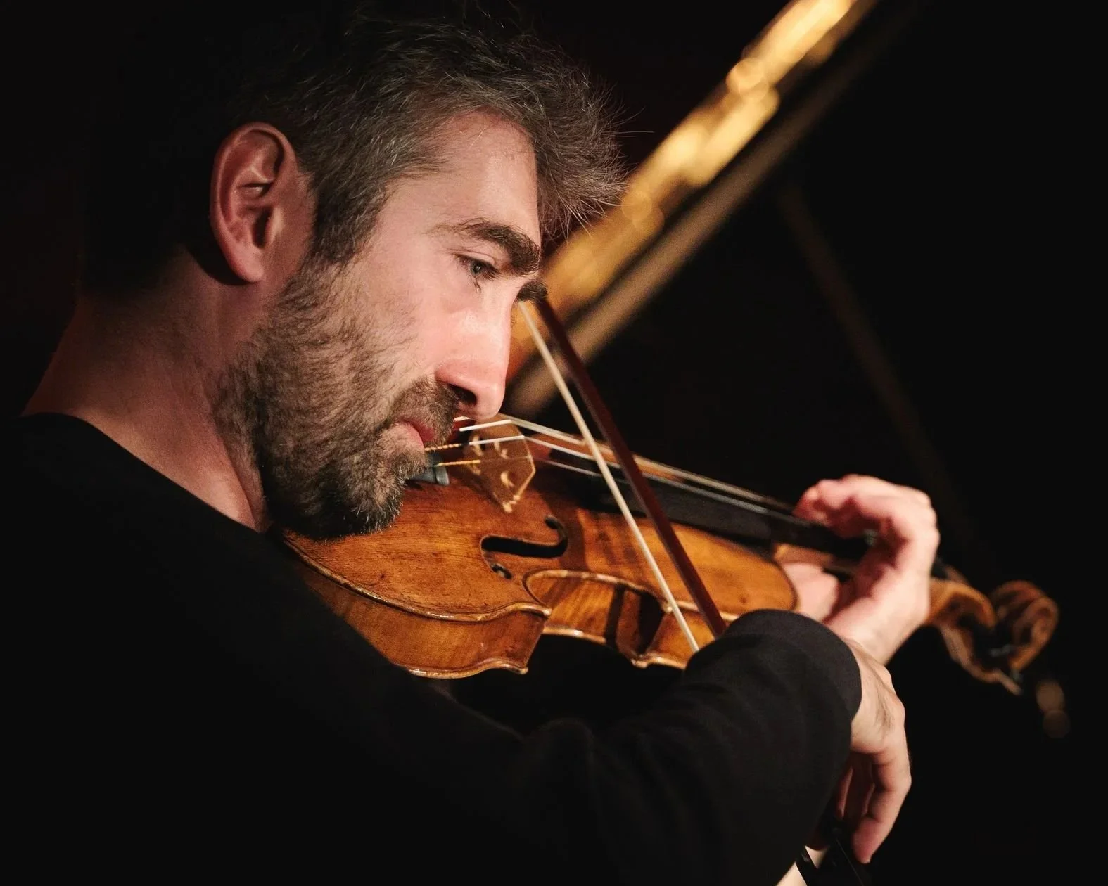 Un homme joue du violon dans une atmosphère sombre. Il a une barbe grise et porte une chemise noire. La scène est en gros plan, concentrée sur son visage et son instrument.