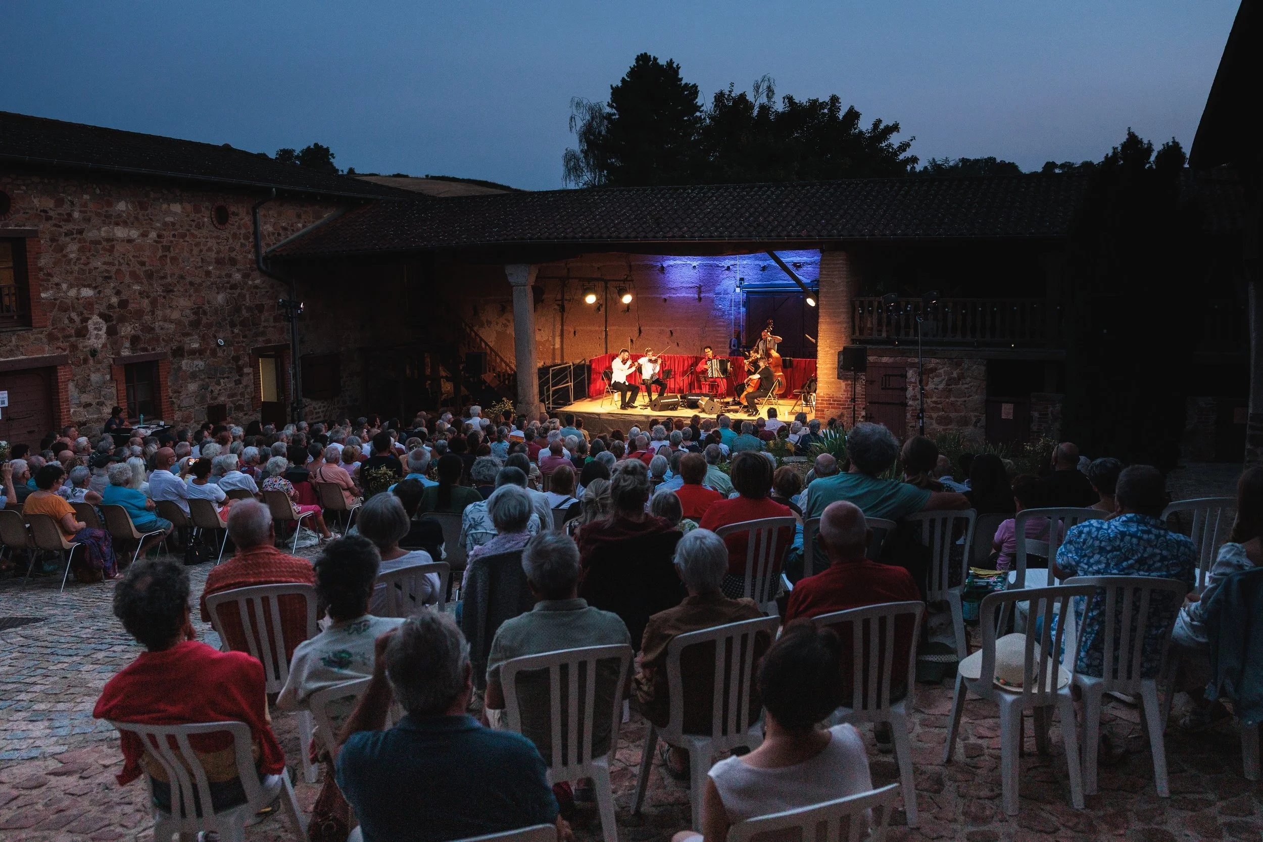 Un concert en plein air se déroule dans une cour en pierre, avec un groupe de musiciens jouant sur scène sous un éclairage. La foule assise regarde la performance lors d'une soirée en été.