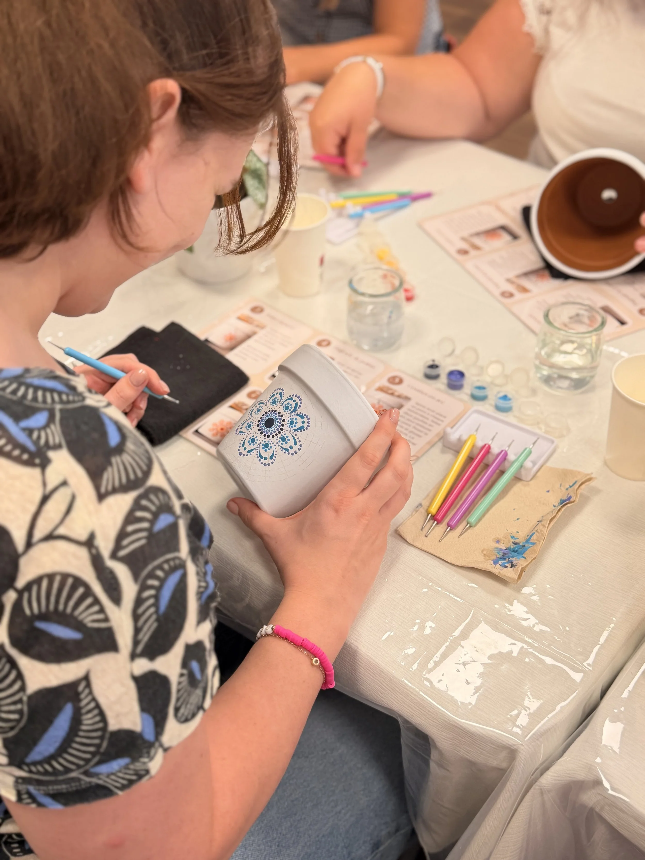 A person with reddish-brown hair and a patterned shirt holding a small decorated box, sitting at a table with art supplies, including colored pens, paint, and printed instructions, during a crafting activity.