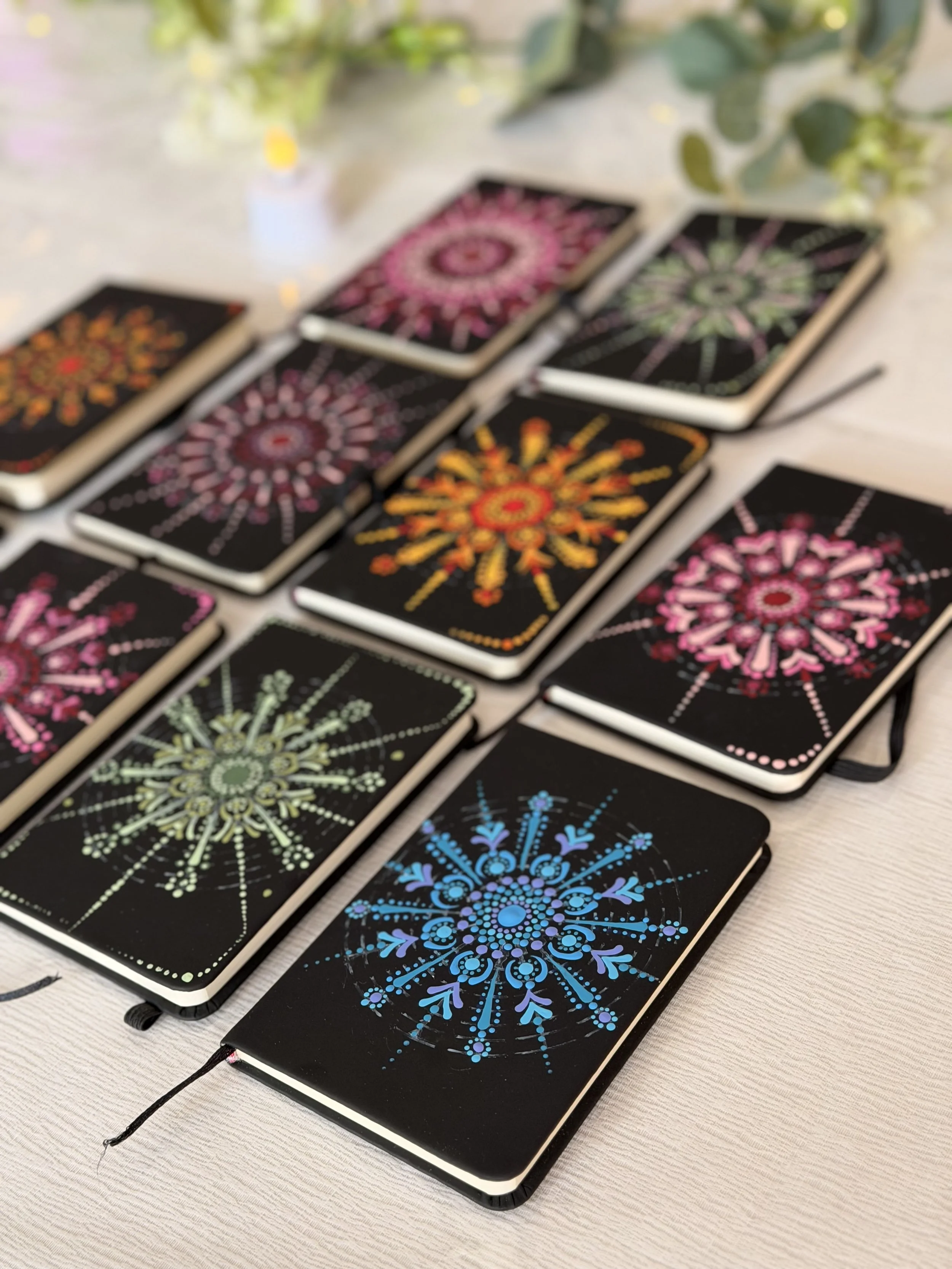 Multiple notebooks with colorful geometric mandalas designs on their covers, laid out on a table with some plants in the background.