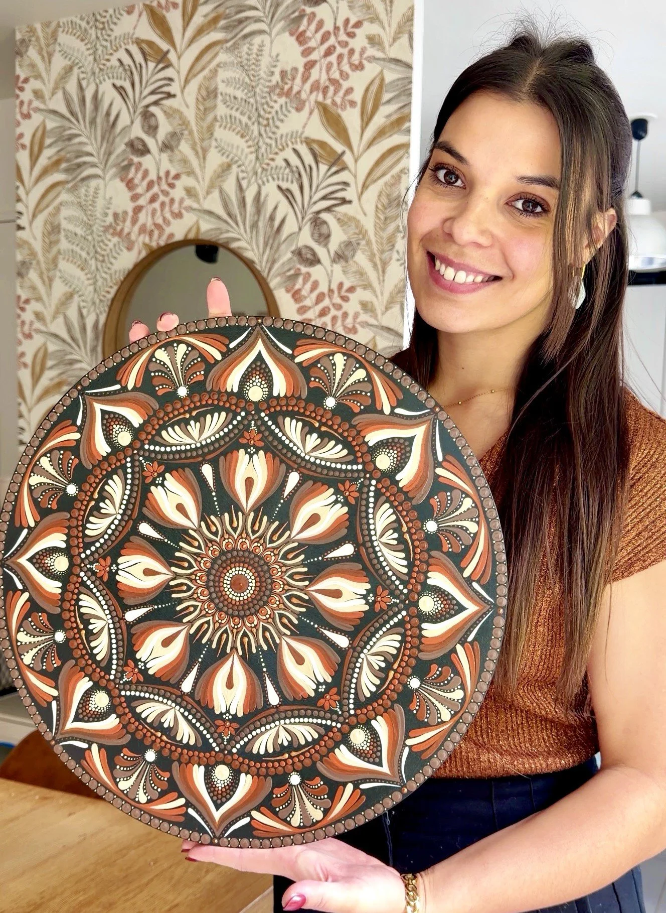 A woman with long brown hair smiling and holding a large, round, elaborately patterned artwork with black, orange, and white colors. The background features wallpaper with a leafy botanical design.
