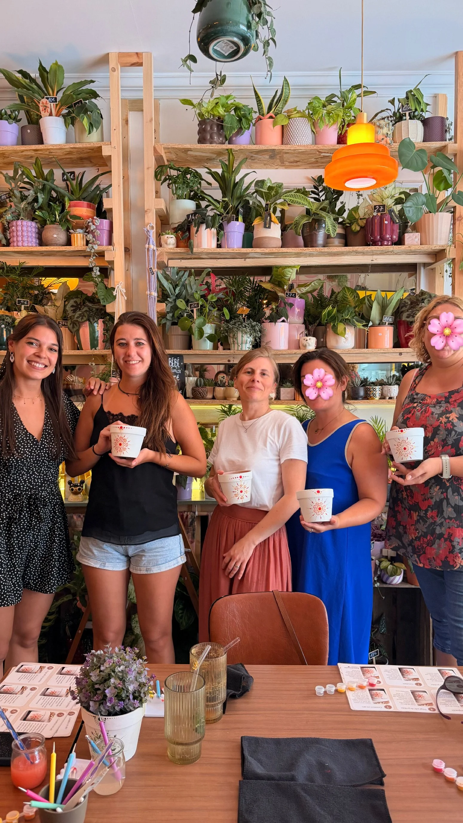 Four women standing together in a plant-filled cafe or shop, holding cups and smiling. There are various potted plants on shelves behind them, some with small chalkboard signs, bright orange and pastel-colored lighting fixtures, a wooden table with test kits, drinks, and pens in the foreground.
