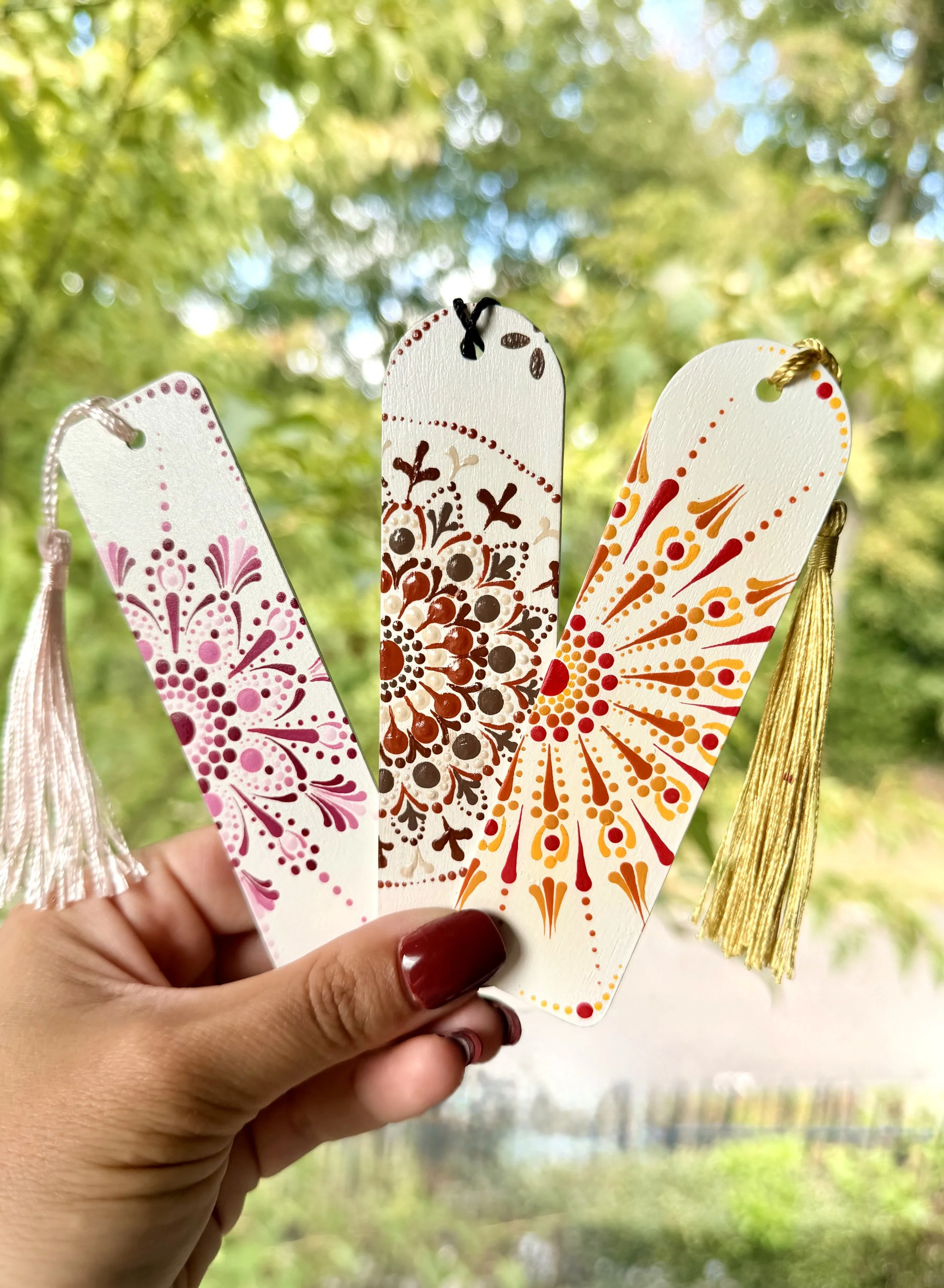 Close-up of three colorful wood bookmarks with decorative patterns being held by a person with painted nails, with a blurred outdoor background of trees and water.