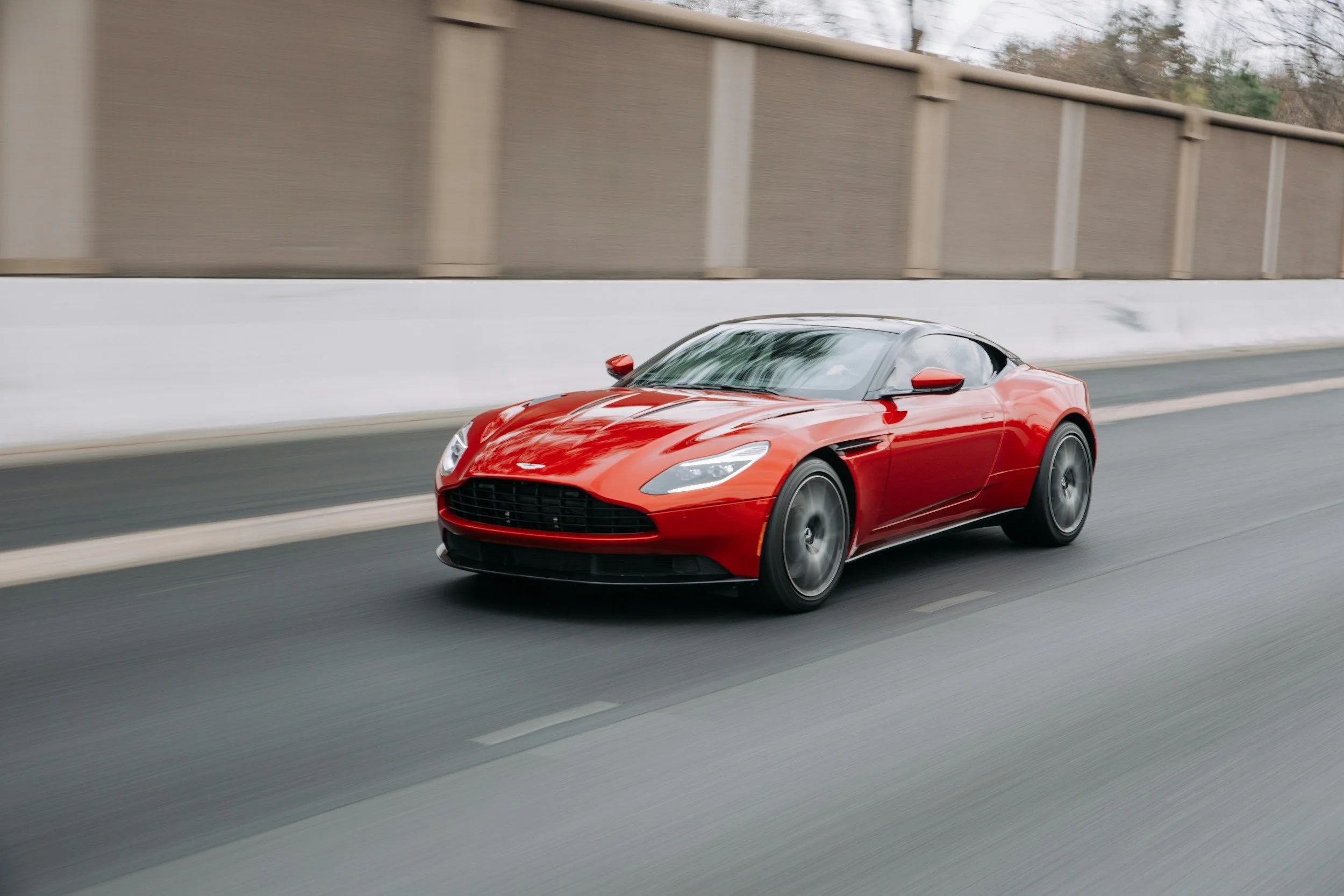 A red convertible sports car driving on a highway next to a tall concrete wall with trees in the background.